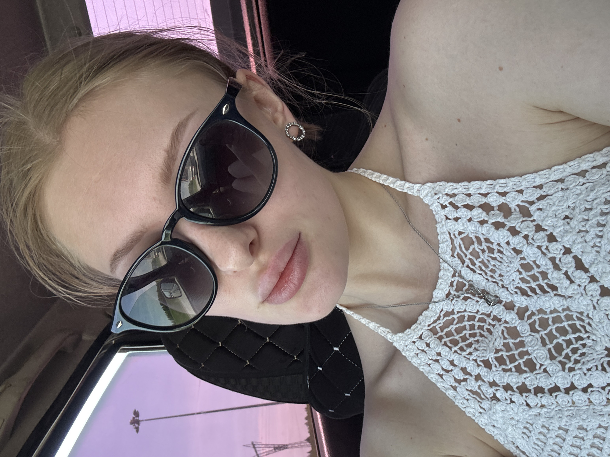A selfie image depicts a woman with light skin and blonde hair, wearing sunglasses and a white crocheted top. She has a silver necklace with a pendant. The background shows the interior of a vehicle with black quilted seats and a pink-tinted window displaying an exterior view of pylons or utility poles. Based on the information and visual evidence, the location is likely the city of Perm, Russia.