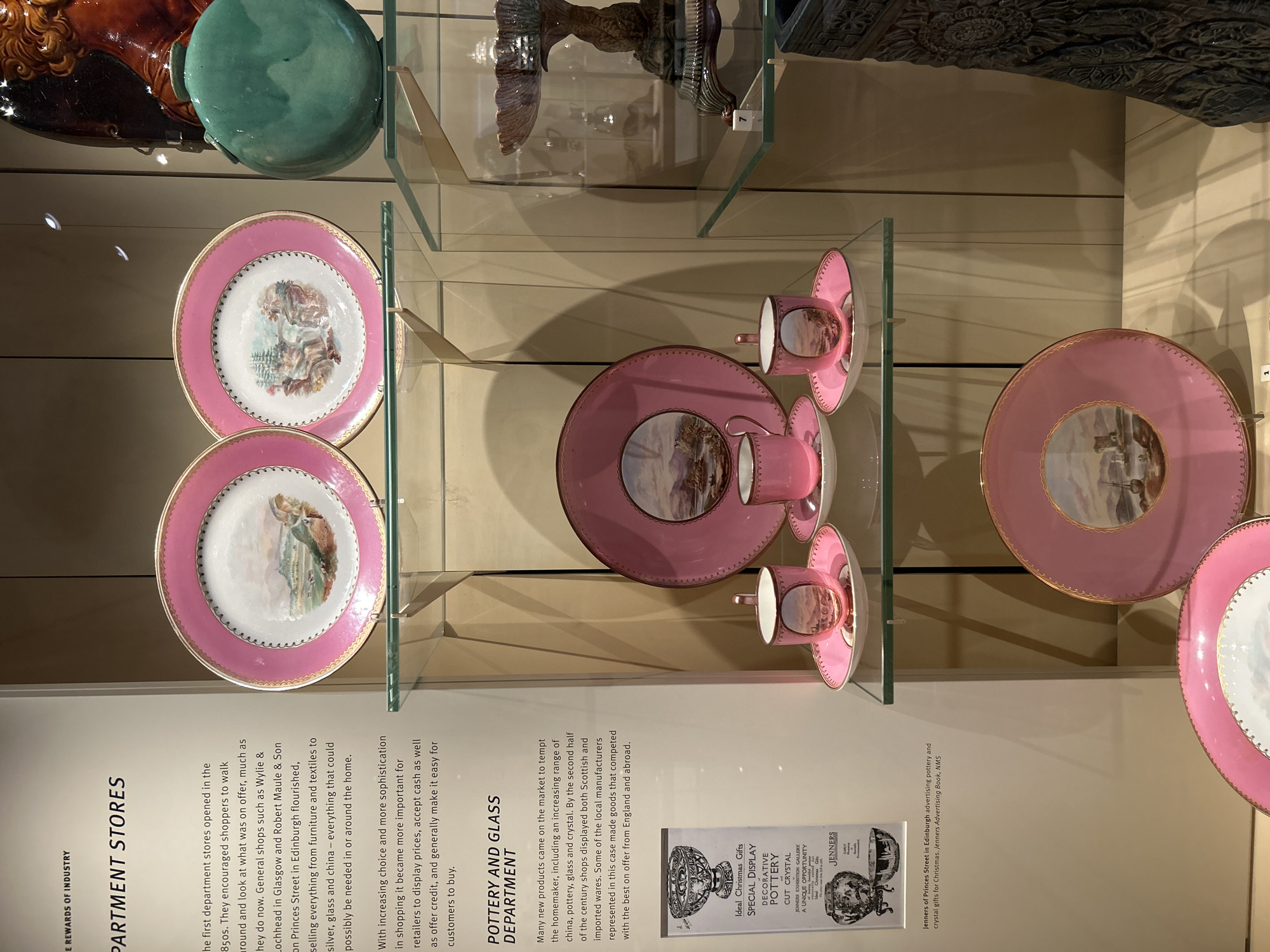 The image shows a museum exhibit featuring a display of pottery and glass items. The text on a panel to the left indicates that the exhibit is about "Department Stores" and the "Pottery and Glass Department". The text also mentions that the first department stores opened in the 1850s and that retailers encouraged shoppers to browse. It also mentions Wylie & Lochhead in Glasgow and Robert Maule & Son on Princes Street in Edinburgh as examples of early department stores. The display includes several pink plates with decorative painted scenes, as well as teacups and saucers. There is also a close-up image of a Jenner's advertisement for pottery and crystal gifts, identifying the location as Princes Street in Edinburgh. The background appears to be a wall with vertical paneling, and the display items are placed on glass shelves. The lighting in the scene creates reflections on the glass surfaces.