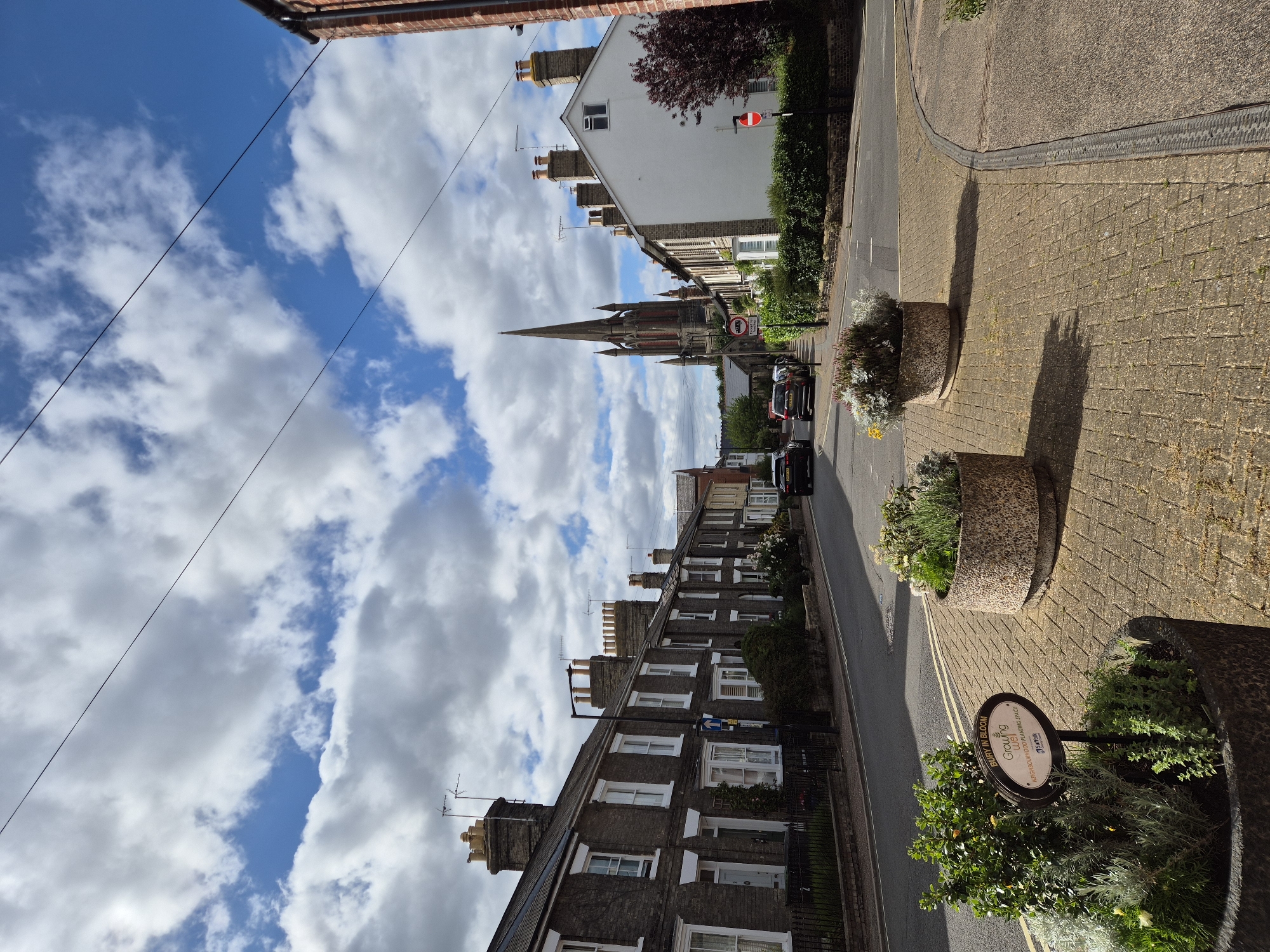 The image depicts a street scene in Bury St Edmunds, West Suffolk, United Kingdom. On the left, a row of terraced houses with dark brick facades and white-framed windows stretches along the street. Several of these houses have chimneys and aerials on their roofs. A black car is parked on the street in front of the houses, and another black car is driving away from the camera.
In the center of the image, a tall, spired church building dominates the skyline, with intricate architectural details. Adjacent to the church, on the right side of the street, is a white building with a red-tiled roof. A dark red tree with dense foliage stands next to this building.
The street is paved with asphalt, and the sidewalk is made of yellow and grey brick pavers. Two large, circular concrete planters containing greenery and flowers are positioned on the sidewalk in the foreground. One planter has a sign attached to it that reads "BURY IN BLOOM Growing well NEIGHBOURHOOD PLANTING SPACE Suffolk".
The sky is partly cloudy, with a mix of white and grey clouds against a bright blue background. Power lines are visible crisscrossing the sky. Several traffic signs are present on the street, including a blue directional sign and two red circular signs with white bars, indicating prohibitions. The overall atmosphere is that of a residential street on a bright, possibly sunny day.