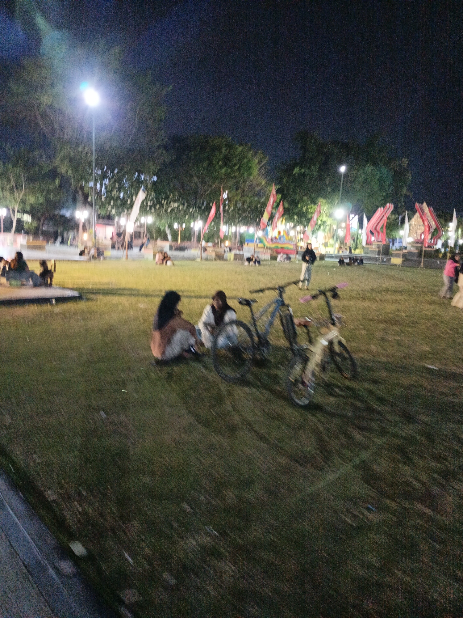 The image depicts an outdoor scene at night in a park-like setting, likely in Klaten, Indonesia, given the context. A large, grassy area dominates the foreground, with several people seated or standing in the mid-ground and background. Three bicycles are present in the foreground, one of which is partially obscured. In the mid-ground, two individuals are seated on the grass, facing away from the camera. Further back, other individuals can be seen gathered. The background features trees, lampposts, and what appear to be decorative structures or flags, illuminated by artificial lighting. The overall atmosphere is relaxed, suggesting a public gathering or leisurely evening activity. The lighting conditions are low, resulting in some motion blur throughout the image.