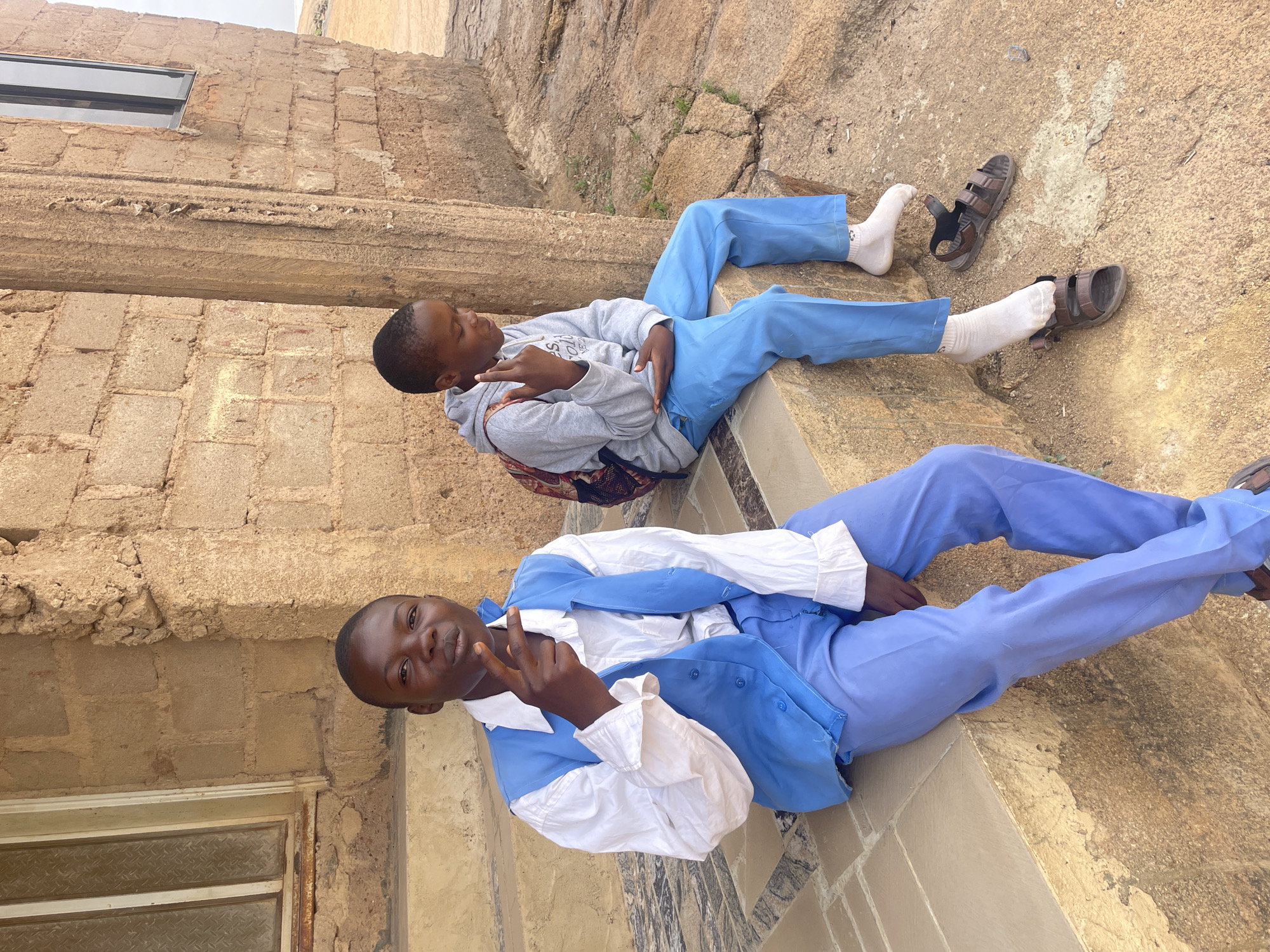 Two young men are resting on the steps of a light brown stone building. The foreground figure wears a light blue uniform jacket over a white shirt and matching light blue trousers. He is gesturing with his fingers. Behind him, a second young man wears a grey hooded sweatshirt, matching light blue trousers, and one sock. His brown sandals sit beside him. The stone building appears weathered, with various crevices and textures. A window can be seen in the building behind the two men. The location is likely in the city of Jos, Nigeria.