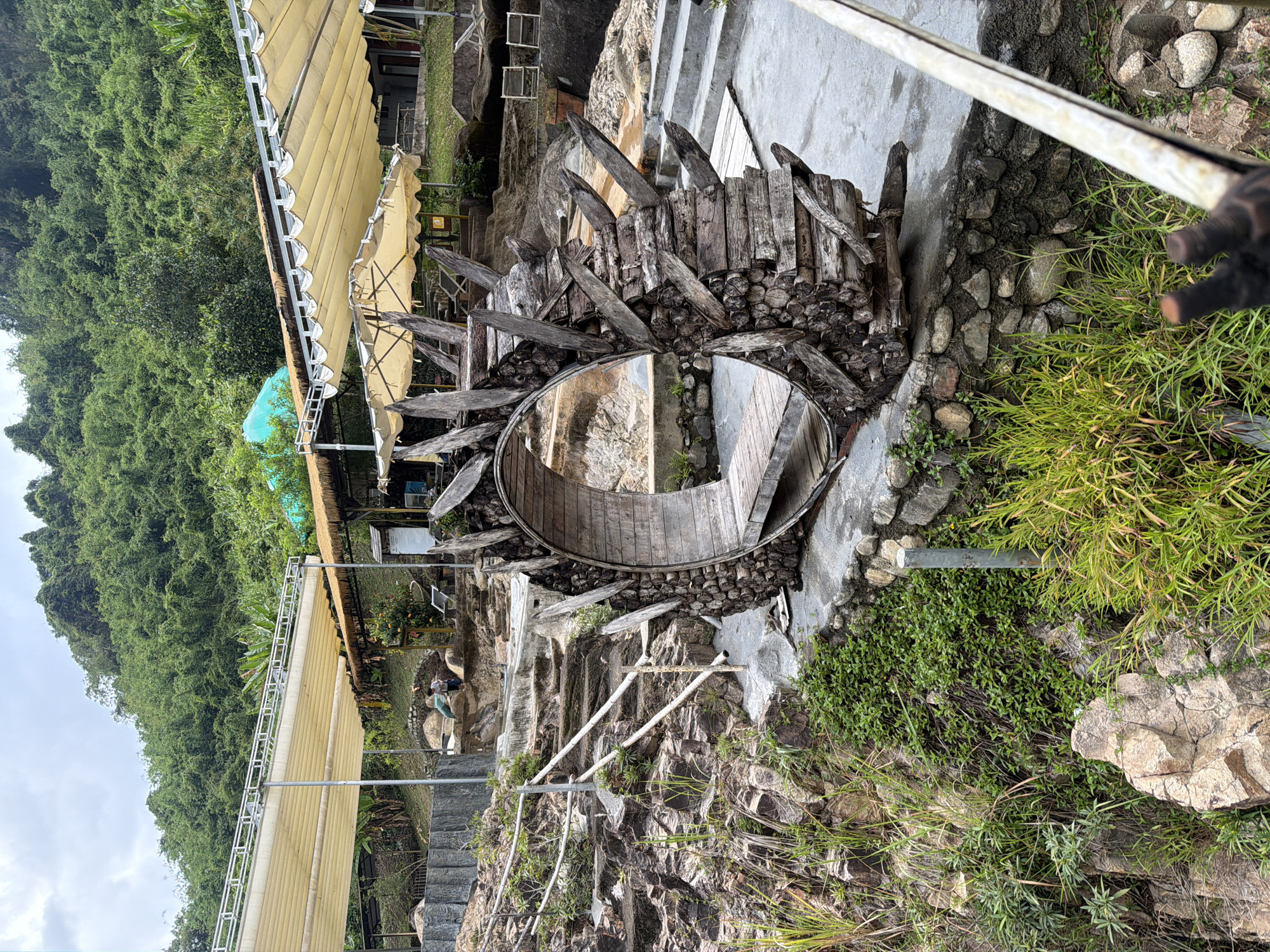 The image shows a rustic outdoor seating area with a distinctive circular wooden structure in the foreground. This structure, made of stacked logs and branches with pointed wooden extensions, resembles a wheel or a natural archway and has a wooden bench inside. The area appears to be a resort or park, set against a lush, green mountainside. In the background, there are several canvas awnings providing shade, some tables, and chairs, suggesting a place for relaxation. Stone steps and rocky terrain are visible, indicating a natural, uneven landscape. Two people are partially visible in the mid-ground, near the steps, interacting with the environment. The overall scene is outdoors, with vegetation and natural rock formations dominating the landscape.