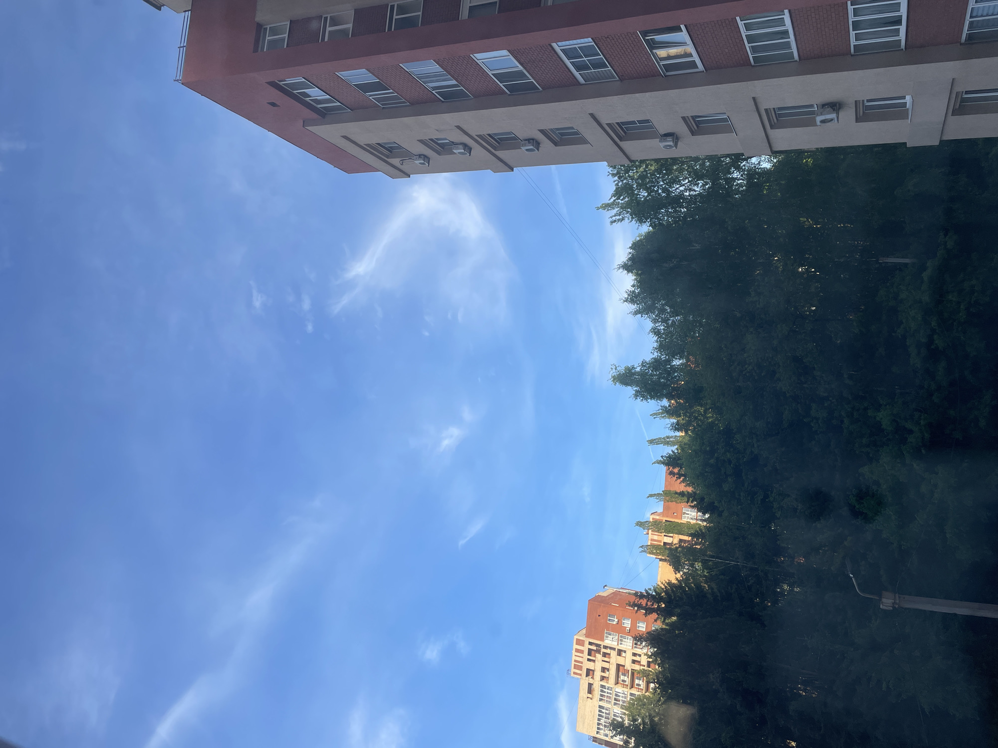The image is a low-angle shot of buildings, trees, and a blue sky with light wispy clouds in Ufa, Russia.
A multi-story building with a red and beige exterior is partially visible at the top of the frame. The building has several windows with white frames. There are also air conditioning units attached to the building.
Dense green trees are present on the right side of the image, partially obscuring another multi-story building with a similar color scheme, visible near the bottom of the frame.
A streetlight pole is slightly visible on the lower right. The rest of the view is clear blue sky.