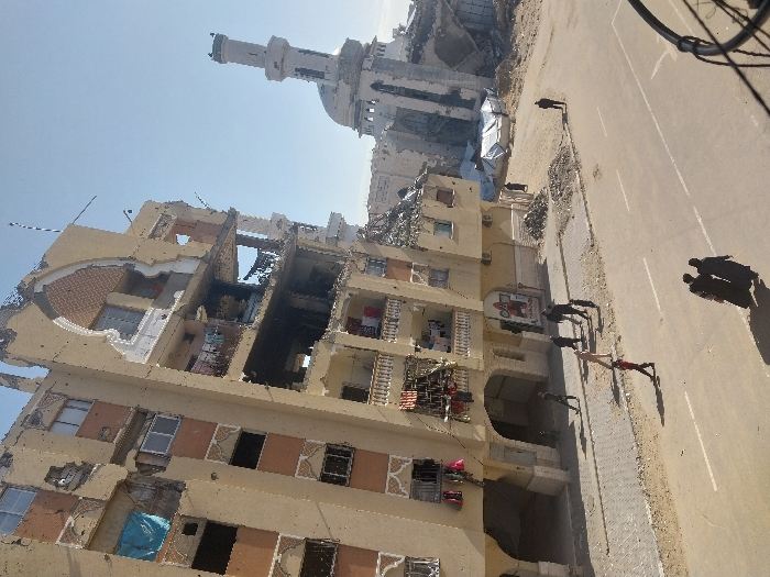 An urban scene in Khan Yunis, Palestinian Territory, depicts significant structural damage under a clear blue sky. In the foreground, a two-lane asphalt road with faded white markings stretches horizontally across the lower third of the frame. Two individuals, dressed in dark, long garments, walk away from the viewer on the road's right side, casting long shadows.
To the left, a multi-story residential building, appearing to be at least seven stories tall, exhibits severe damage. Its light-colored facade is heavily pockmarked, with large sections of walls missing, exposing interior rooms and structural rebar. Multiple windows are shattered or absent, and some balconies are damaged, though laundry is visible hanging from several upper-level balconies. A large decorative arch feature is prominent on the upper floors. Below, several people, including a child in a red shirt, walk along the paved sidewalk and street in front of the damaged building. Piles of dirt and rubble line the sidewalk base.
In the mid-ground to the right, a mosque with a distinct minaret and dome is visible, also showing signs of damage and partial collapse, with debris surrounding its base. Beyond the mosque, mounds of rubble and at least two white tarp-covered structures are present. A lone individual walks towards this rubble-strewn area. The overall environment is one of extensive destruction, with dust and debris covering parts of the ground, yet with visible human movement through the area.