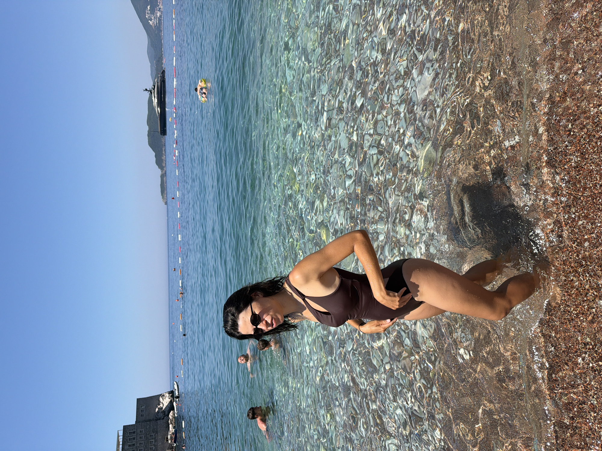 A woman stands in shallow, clear water on a pebbly beach. She is wearing a dark brown one-piece swimsuit and sunglasses. Her hair is dark and wet. In the background, other people are in the water, and a large yacht is visible on the horizon. The water is turquoise and the sky is clear blue. The location is the beach in the city of Rađenovići, Montenegro.