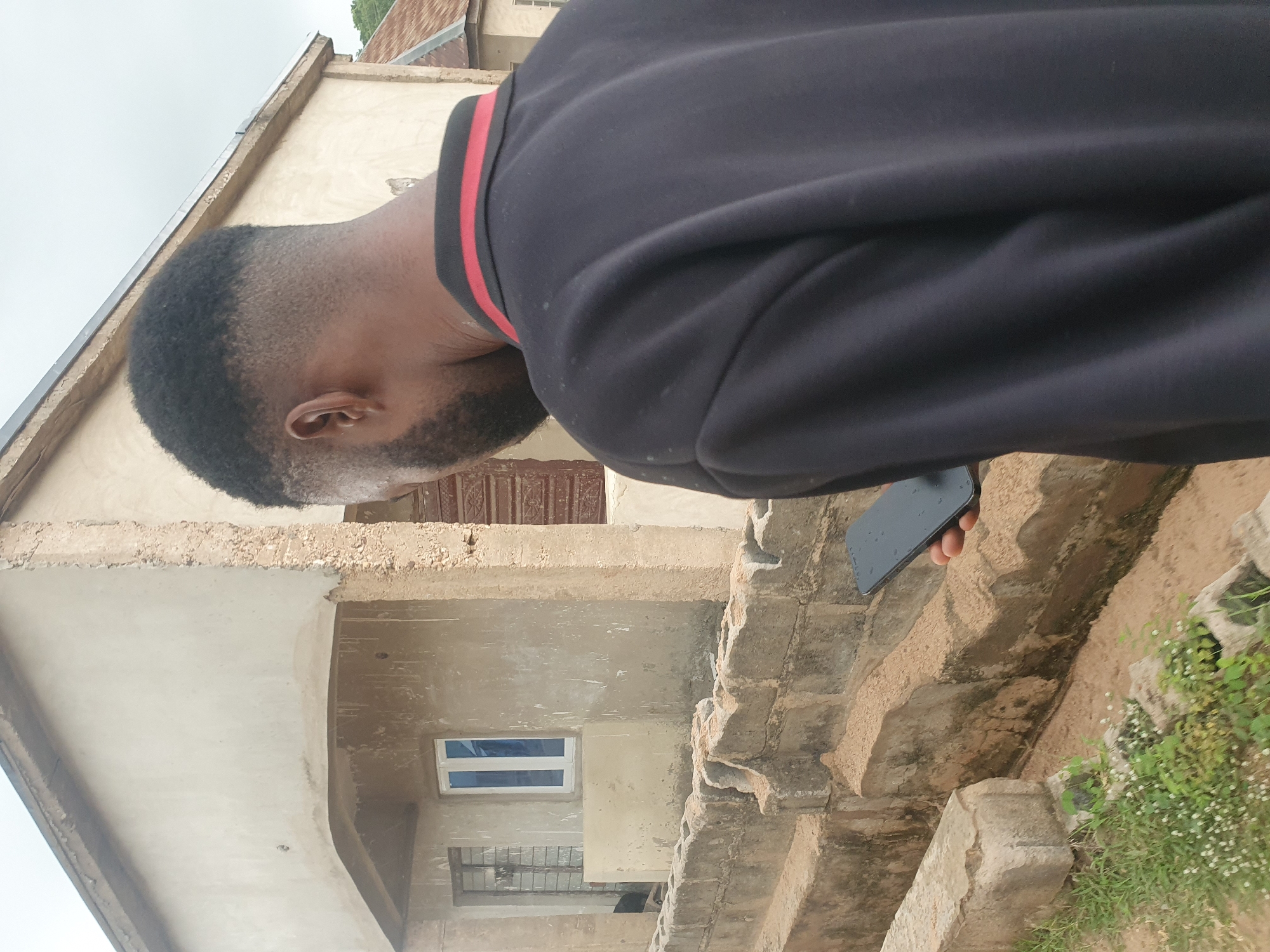 A person, viewed from behind and from the side, stands outdoors in what appears to be an unfinished or partially constructed residential area. The person is wearing a dark long-sleeved shirt with red and black trim around the collar. Their right hand holds a dark-colored smartphone.
The background features a building with rough, unpainted concrete walls, exhibiting signs of construction. One window is visible, with a white frame and blue panes. In front of the building, a low wall constructed from concrete blocks runs horizontally across the mid-ground. The ground is uneven and appears to be a mix of dirt and sand, with some sparse green vegetation and small white flowers in the lower left corner. The overall impression is of a developing or perhaps neglected urban or semi-urban environment in Lokoja, Nigeria. The sky is overcast.