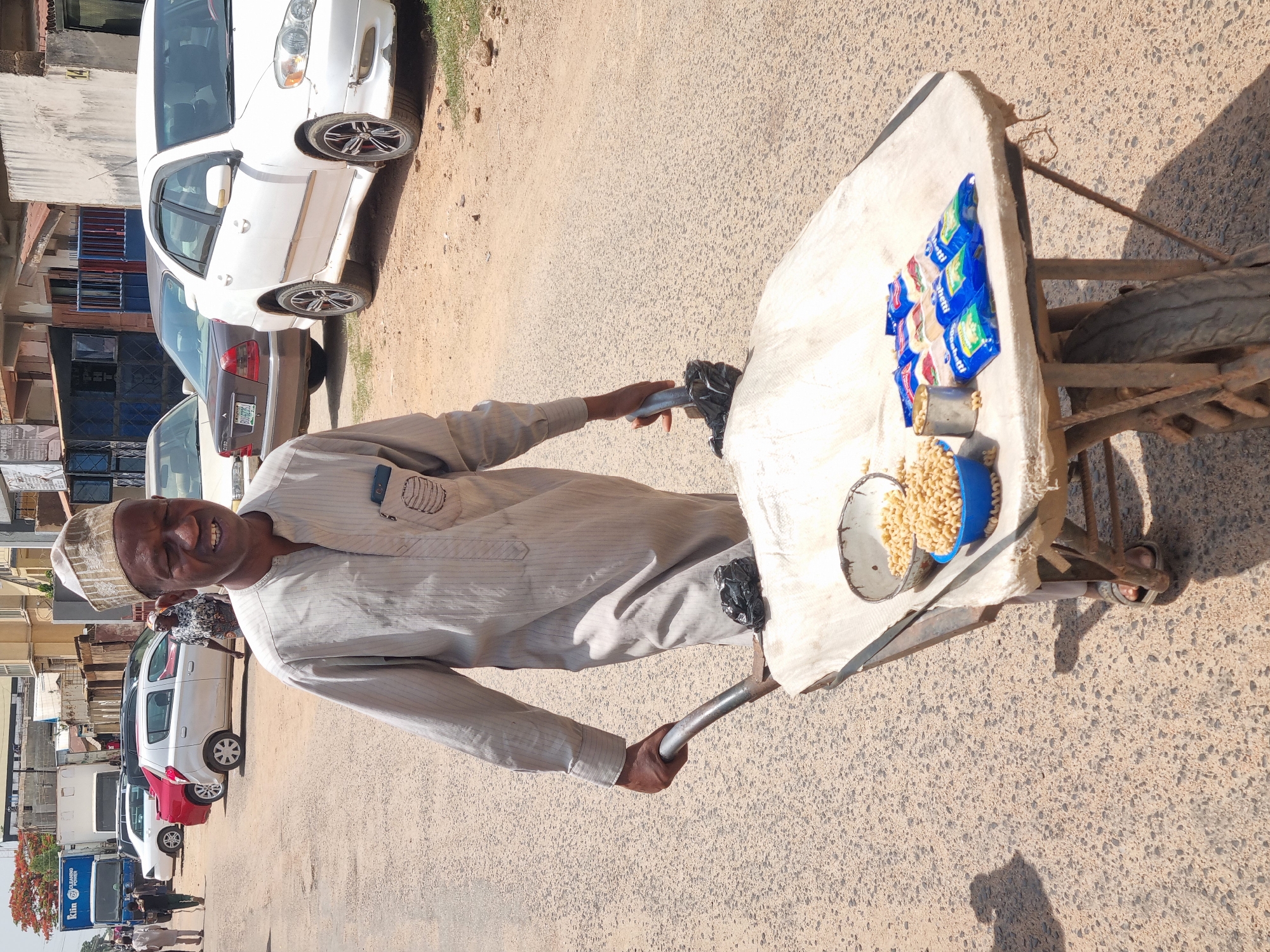 A man, likely a street vendor in Jos, Nigeria, stands next to a makeshift cart. He is smiling, wearing a light-colored, long-sleeved shirt and a white cap. The cart has a white surface displaying small blue bags of pasta and a container of loose pasta. The cart is wheeled and appears to be made of wood and metal. He is holding onto the cart's handles. In the background, there are cars parked along a street lined with buildings. The ground is a light tan color, possibly asphalt or concrete. Several people are walking in the background, near some trucks and vehicles.