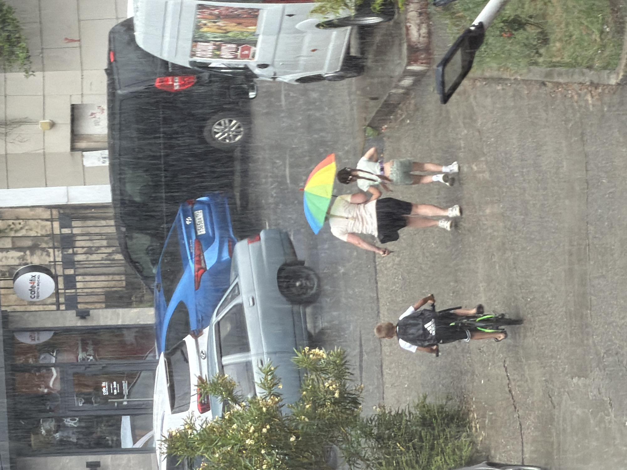 In Sochi, Russia, during a rainstorm, a young boy with a black backpack rides a bicycle on a wet asphalt path. Behind him, an adult and a child walk together under a multicolored umbrella. Several vehicles, including a white van and a dark minivan, are parked nearby, and a light-colored sedan is visible further in the background. The scene unfolds in front of buildings with storefronts, one of which is identified as "cafe4fix." Green foliage from a tree with light-colored flowers is present in the foreground, contributing to the natural elements of the urban environment.