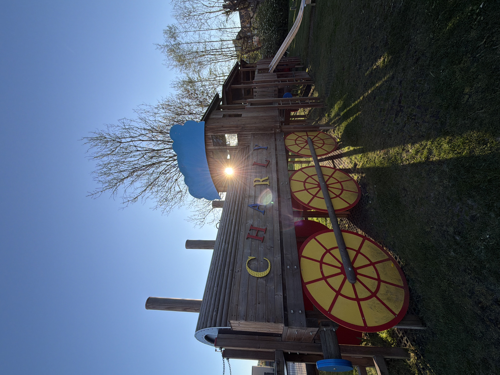 The image displays an outdoor playground scene in Niederanven, Luxembourg, under a clear, bright blue sky. The central subject is a large wooden play structure designed to resemble a train, complete with a front boiler section and multiple carriage-like segments. No people are present, giving the scene a serene atmosphere.
The wooden structure is detailed with six prominent, decorative yellow wheels featuring red spoke patterns, three on each side. A distinctive blue, wave-shaped element is attached to the front of the train structure. Along the side of the train, the name "CHARLY" is spelled out in individual, colorful block letters: a yellow 'C', orange 'H', blue 'A', red 'R', yellow 'L', and orange 'Y'.
In the background, trees with mostly bare branches suggest an early spring or late autumn setting. A silver slide is visible on the far side of the wooden structure, alongside various ladders and platforms, confirming its use as a children's play area. The sun is low in the sky, creating a strong lens flare that partially brightens the train's side and casts long shadows across the neatly mown green grass, indicating either early morning or late afternoon. The weather is sunny and cloudless.