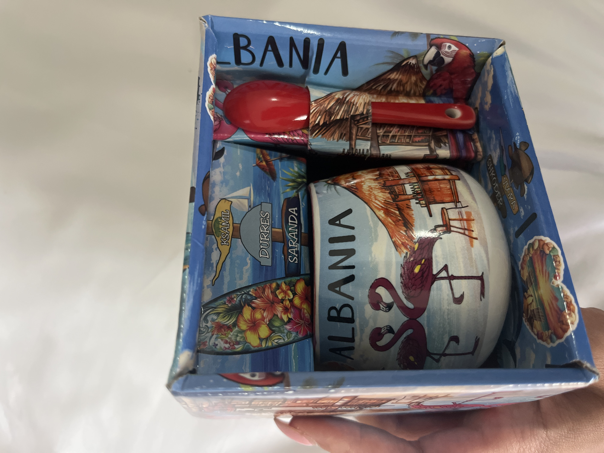 The image shows a souvenir gift box from Albania. The box is blue and decorated with images of tropical scenery, including palm trees, beaches, flamingos, and a parrot. Text on the box includes "ALBANIA", "KSAMIL", "DURRES", and "SARANDA", which are all locations in Albania. Inside the box is a mug, a spoon, and possibly other small items. The mug has an image of two flamingos and a beach bar. The spoon is red. A hand is holding the box, and the background is a plain white surface. The box appears to be made of cardboard.