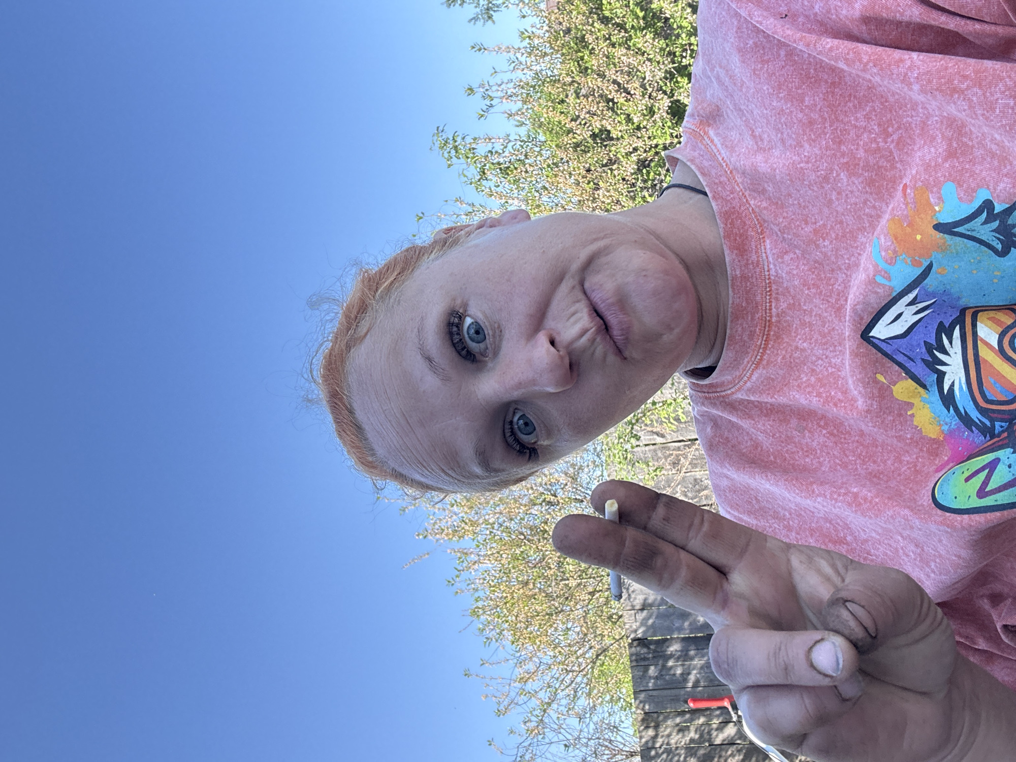 A woman with red hair is holding a cigarette between her fingers, outdoors. The sky is blue and partially visible with a tree in the background. The woman is wearing a coral-colored sweatshirt. The background contains a wooden fence. The location is not identifiable from this image.