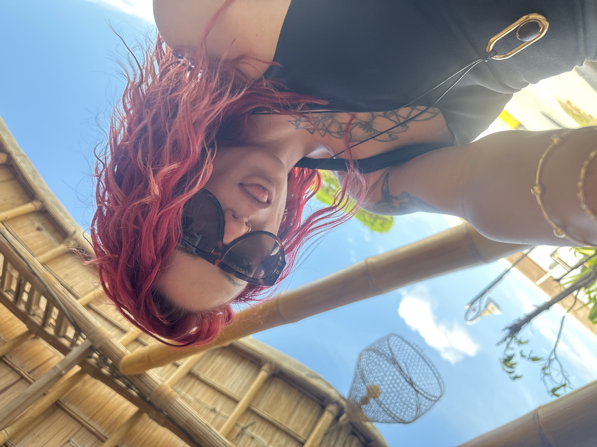 A woman with red curly hair and sunglasses is looking down at the camera with her tongue sticking out. She is wearing a black top and has tattoos on her chest and arm. She is indoors with bamboo structures forming a roof. Decorative light fixtures hang from the ceiling. The sky is visible through the structures. The location is Unknown, Indonesia.