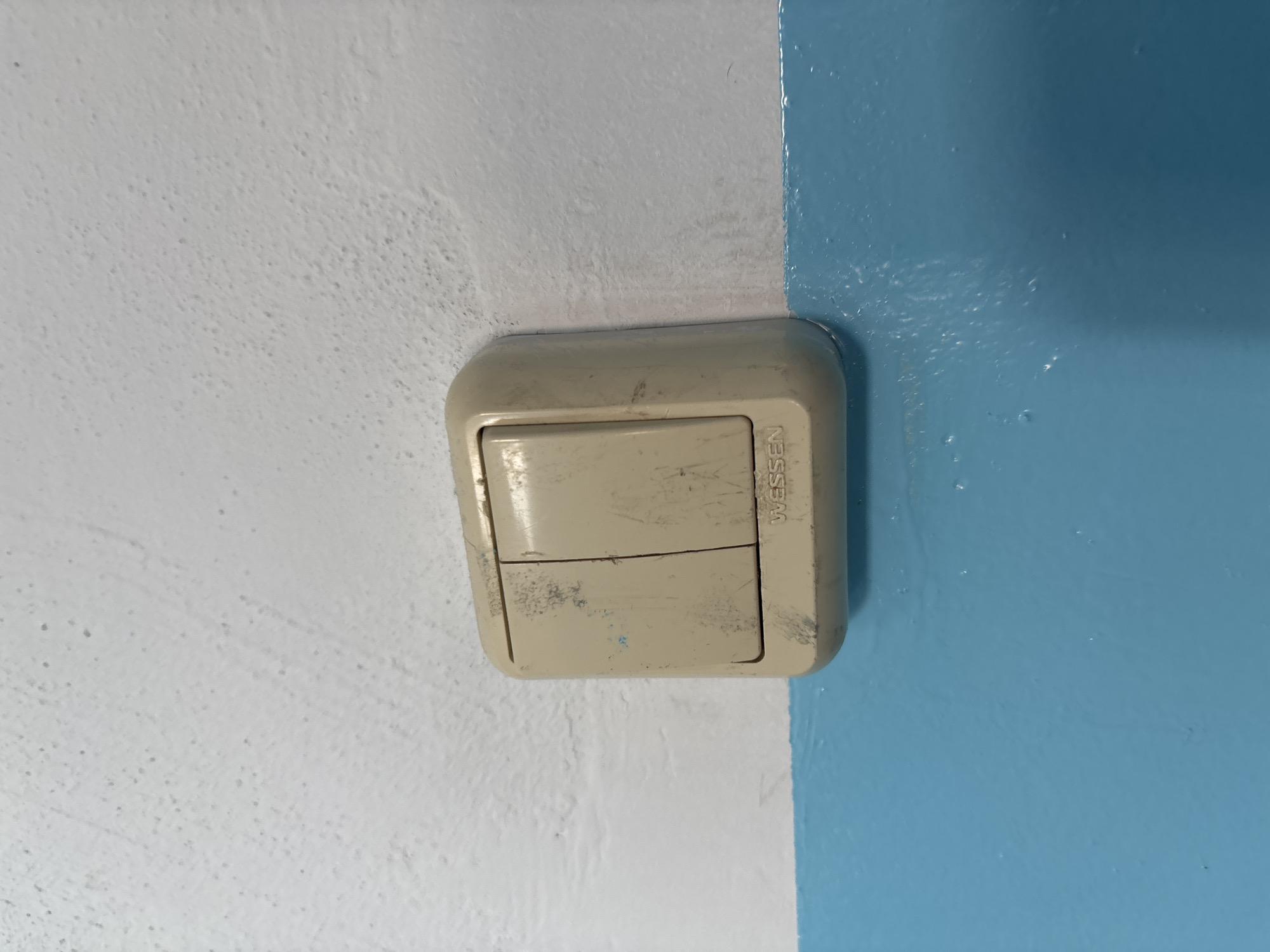 The image shows a close-up of a beige, two-gang light switch with the brand name "WESSEN" visible. The switch is mounted on a wall that is painted white on the top half and a bright blue on the bottom half. The white portion of the wall has a textured, slightly speckled appearance, while the blue portion is smoother and appears to be a glossier finish. The light switch itself appears worn, with scuff marks and some blue paint transfer on its surface, suggesting it may have been exposed to painting work or general wear and tear. There are no people or any other objects clearly visible in the frame, and the image focuses solely on the light switch and the wall it is mounted on. The scene suggests an interior setting, likely a domestic or commercial space. No specific location in Blagoveshchensk, Russia can be identified from this image alone.