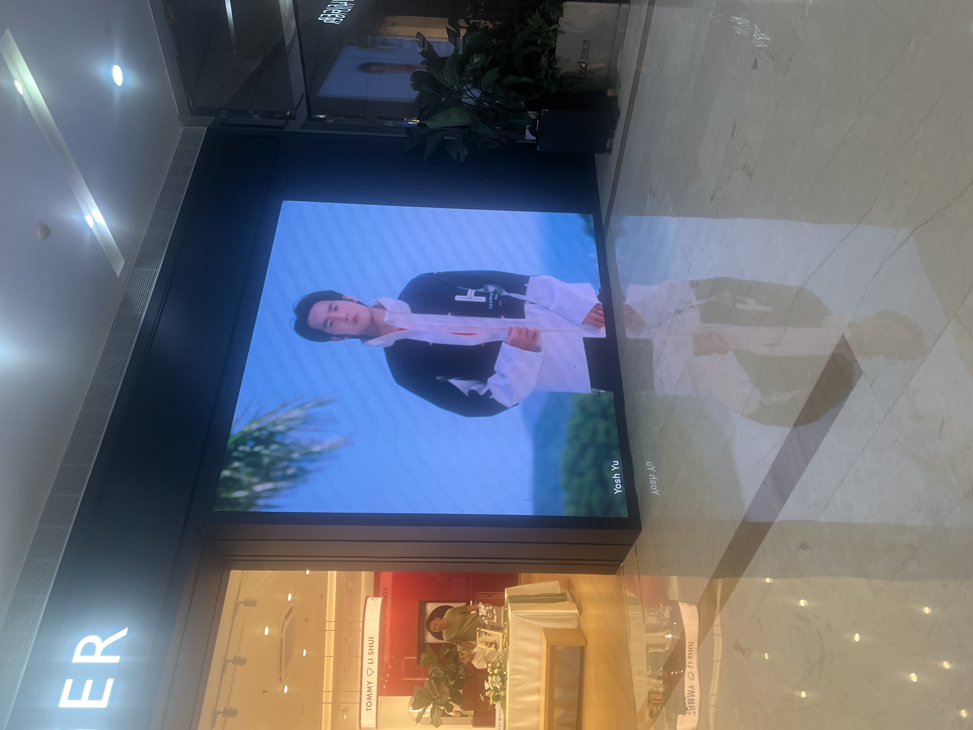 In the Liandu District of China, a digital billboard displays a young man wearing a black and white jacket with the letter 'H' prominently featured. The billboard is situated within a retail environment, indicated by a store entrance to the left with a sign that reads "TOMMY ♡ LI SHUI." Inside the store, two individuals are visible near a table adorned with flowers and other display items. The floor reflects the billboard and store lighting, creating a polished and somewhat diffused image. The overall scene appears to be a commercial space within a shopping area.