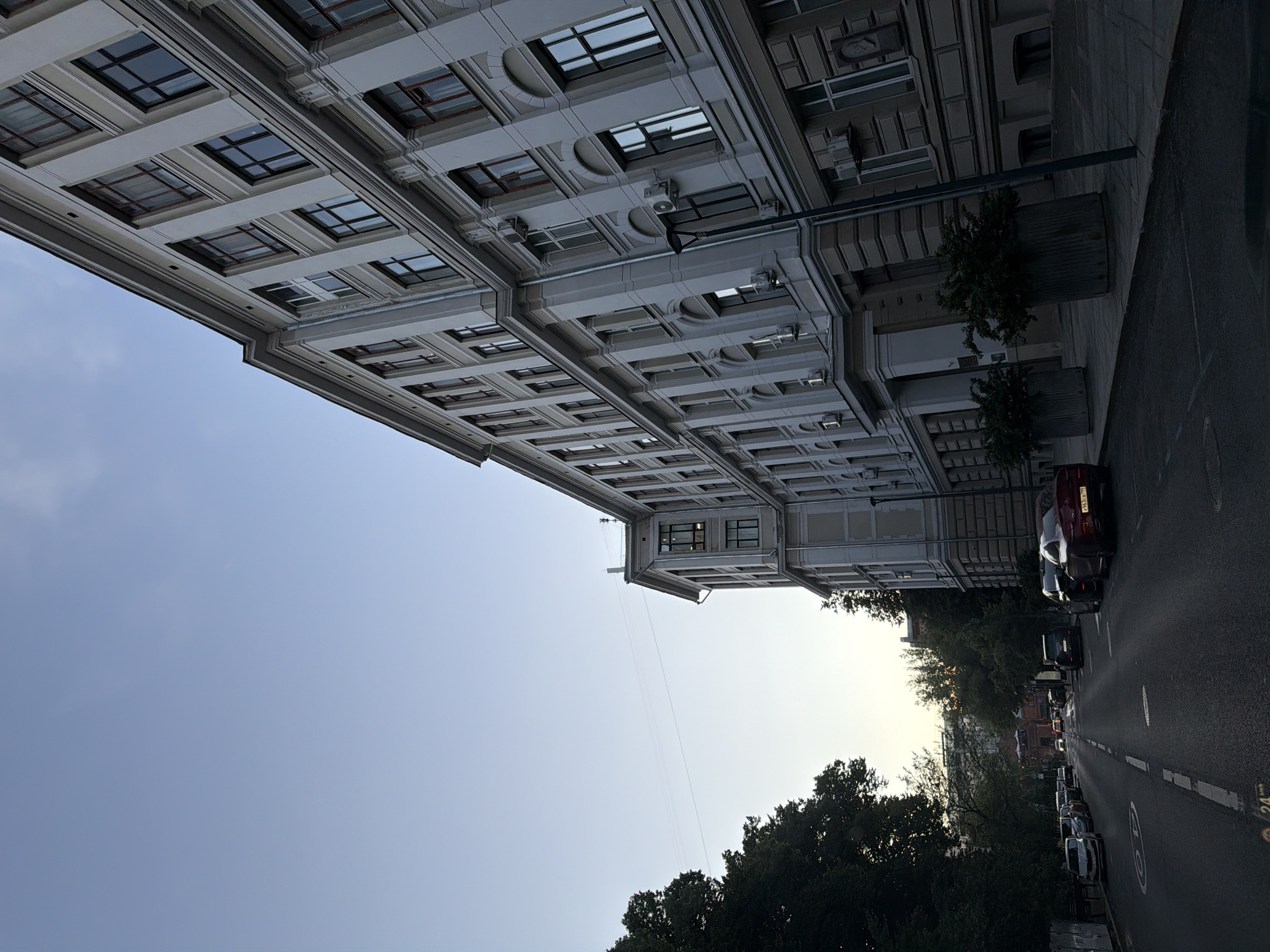 This image depicts a street scene in Moscow, Russia, likely during the evening or early morning based on the lighting. The dominant element is a large, multi-story building with a classical architectural style, characterized by its light-colored facade, numerous windows with dark frames, and decorative detailing. The building stretches along the right side of the frame, angled away from the viewer. In front of the building, there are large planter pots with dark green foliage.
To the left of the building, a street runs parallel to it. Several cars are parked along the side of the street, and others are driving on the road. The street surface appears wet, suggesting recent rain. Trees with dense green foliage line the left side of the street, creating a contrast with the urban architecture. The sky above is a pale blue, with a hint of brightness indicating either sunrise or sunset. A utility pole with a lamp is visible on the sidewalk in front of the building. The overall impression is one of a quiet urban street in a historic district of Moscow.