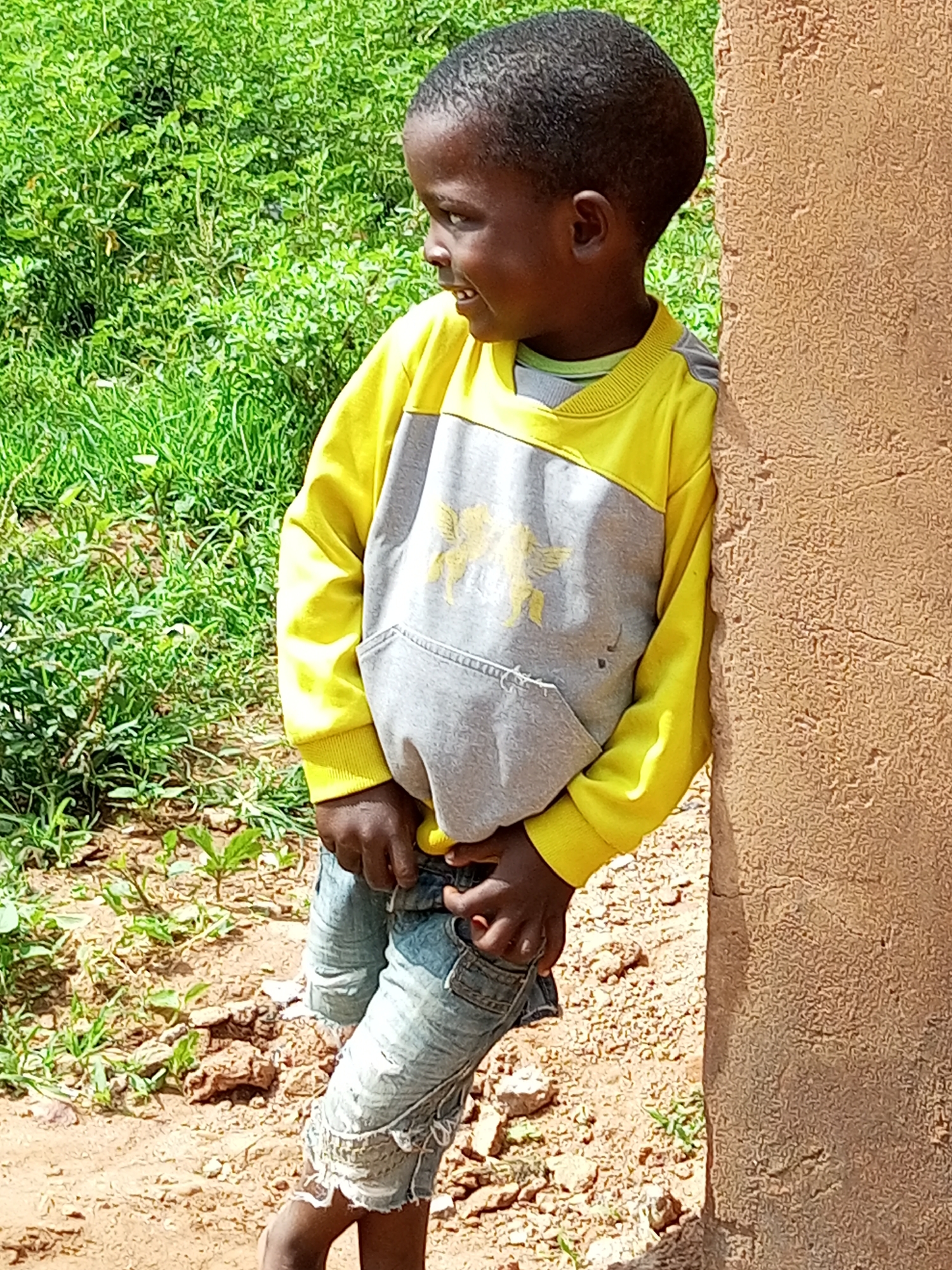 A young boy leans against a textured, light brown wall. He is looking to his left with a slight smile. The boy is wearing a yellow and gray long-sleeved sweatshirt with a graphic design on the front, and distressed denim shorts. He is standing on dirt ground with some sparse green vegetation in the background. The location appears to be an outdoor, informal setting.