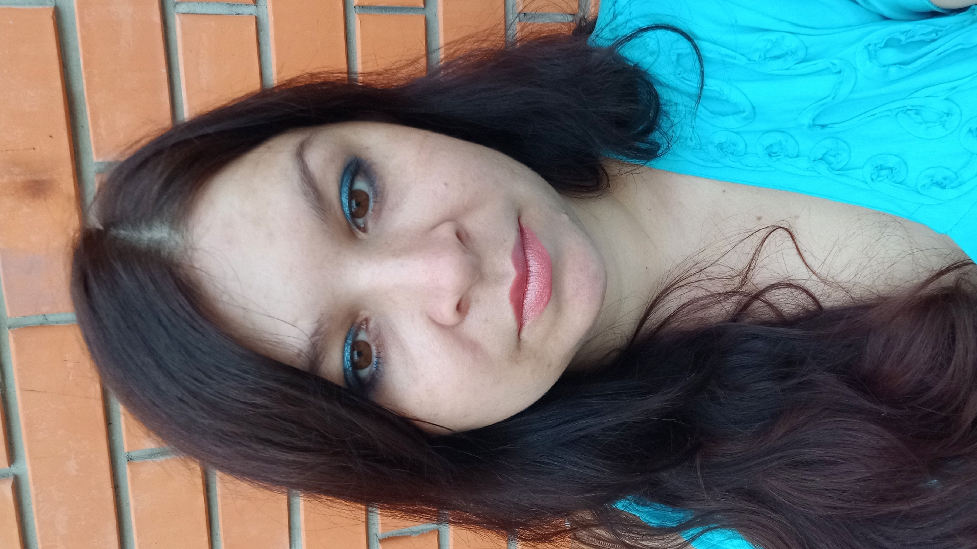 A close-up, medium shot captures a woman with long, dark brown hair and brown eyes, looking directly at the camera. She is wearing a bright turquoise top with a textured design and has applied blue eyeshadow and pink lipstick. The background consists of a brick wall with orange-red bricks and grey grout lines. The setting appears to be outdoors, possibly in the city of Kropyvnytskyi, Ukraine.