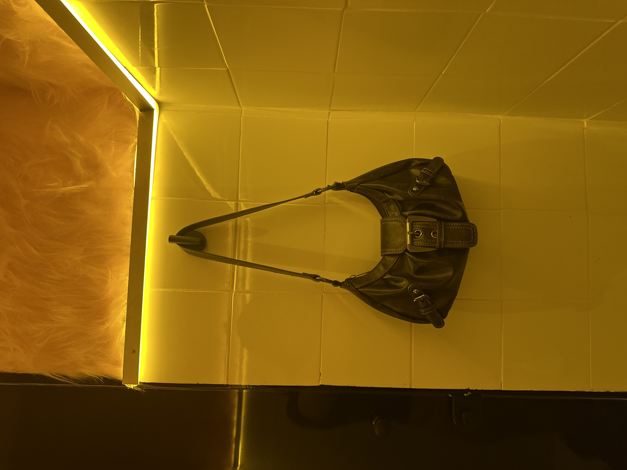 A brown leather purse hangs on a yellow tiled wall with yellow lighting. A fluffy orange surface is seen to the left of the purse, inside an opening illuminated by a strip of yellow light. A dark, glossy surface is at the bottom of the frame. The photograph was taken in Mexico City, Mexico.