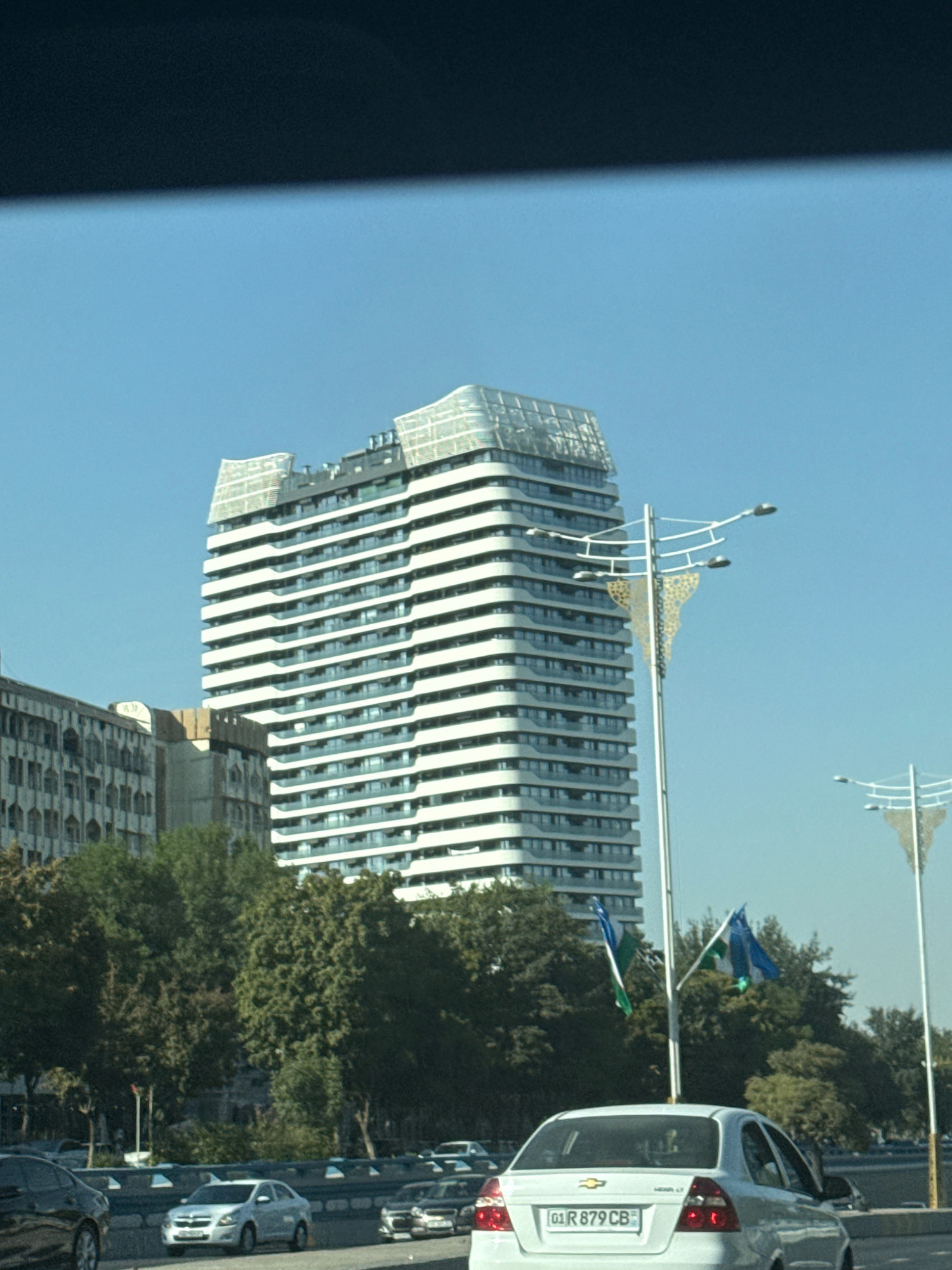 The image shows a daytime exterior scene in Tashkent, Uzbekistan. In the foreground, a white Chevrolet sedan with license plate "01 R 879 CB" is visible, facing away from the camera. The car is on a multi-lane road with other vehicles present, including a silver sedan and a black SUV. The road is bordered by a concrete barrier. Behind the road, a line of mature trees forms a green canopy. To the left of the trees, an older, ornate building with multiple windows is partially visible. Dominating the center and right of the frame is a modern, multi-story building with a distinctive wave-like facade featuring white horizontal banding and reflective glass. Two lampposts with decorative elements are positioned in front of this modern building. Two flags, one predominantly blue and the other green with blue accents, are flying on poles between the lampposts and the modern building. The sky above is clear and blue.