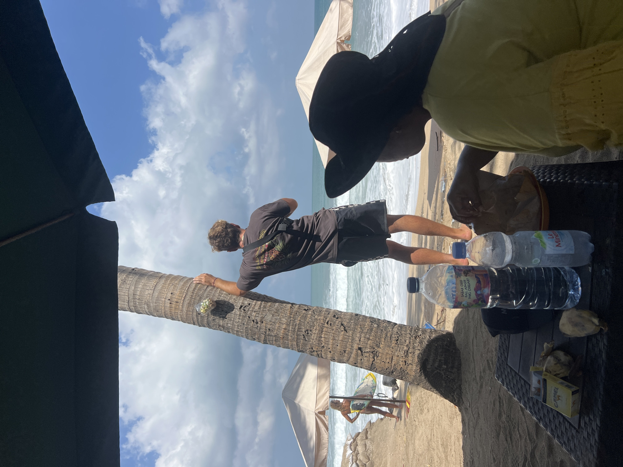 The image shows a beach scene in Kuta, Indonesia. In the foreground, a person wearing a yellow shirt and a wide-brimmed black hat is sitting at a table, holding a piece of food. On the table are a pack of Camel cigarettes, two pieces of food, and two bottles of water. In the mid-ground, a man in a dark t-shirt and shorts stands next to a palm tree, looking out at the ocean. He is holding a clear plastic cup with what appears to be small, green objects inside, and has a red wristband on his left arm. A woman holding a surfboard stands under a beach umbrella in the background. The ocean is visible with waves, and the sky is blue with scattered clouds. The scene is set on a sandy beach.