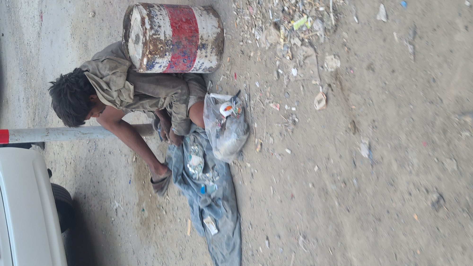A partially obscured, side-view depicts an individual located on the ground next to a white vehicle, adjacent to a dirt ground covered with garbage. The person, dressed in worn, dirty clothing and wearing one slipper, leans against a red and white cylindrical object resembling a barrel. A plastic bag containing items is situated near the person, and clothing lies nearby. The setting is a dirt surface cluttered with various pieces of garbage. The location is in Quetta City Tehsil, Pakistan.