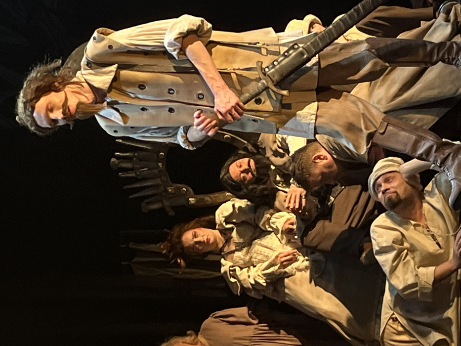 The image depicts a theatrical performance occurring on a stage in a venue located in Novoshakhtinsk, Russia. Six individuals, all adults in period-style costumes, are visible. A tall male figure, characterized by long dark hair, a pronounced handlebar mustache, and a goatee, stands centrally, clad in a light-colored, long-sleeved shirt, a beige waistcoat with dark buttons, and dark knee-high boots. He holds a dark, sheathed sword vertically in his left hand, resting it against his leg, and looks upward and to the right with a serious expression. To his left, a female figure with styled, dark hair and an elaborate light-colored gown with ruffles is seated on a chair, observing forward with a solemn demeanor. Directly behind the seated female, a male figure with dark, curly hair, a full beard, and dark clothing is seated, his hands clasped. To the far left, a partially visible individual in a simple garment is present. In the lower mid-ground, a male is positioned, appearing to be seated or kneeling. In the foreground, another male with a goatee and a light-colored cap is reclining on his back, looking upward. The background is dark, with a noticeable ornate, dark, spiky structure, likely a theatrical prop or part of the set design. Directed stage lighting illuminates the performers.