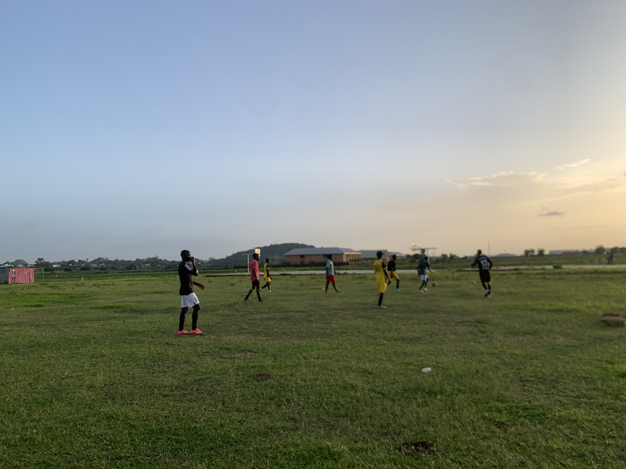 The media file depicts a group of people playing soccer on a grassy field in Gwafan, Nigeria. Several individuals, appearing to be young men, are scattered across the field, some actively engaged in the game, while others are walking or standing. They are dressed in athletic wear, with some wearing brightly colored jerseys. The field is green and appears well-maintained, with some scattered litter. In the background, there are buildings, including what looks like a pink structure with a soccer goal, and a distant tree line. The sky is a clear, pale blue, transitioning to a soft orange and yellow hue towards the horizon, indicating it is either sunrise or sunset. The lighting suggests a peaceful and perhaps cooler part of the day, common for outdoor activities. The overall scene conveys a sense of casual recreation and community engagement in a rural or semi-rural Nigerian setting.