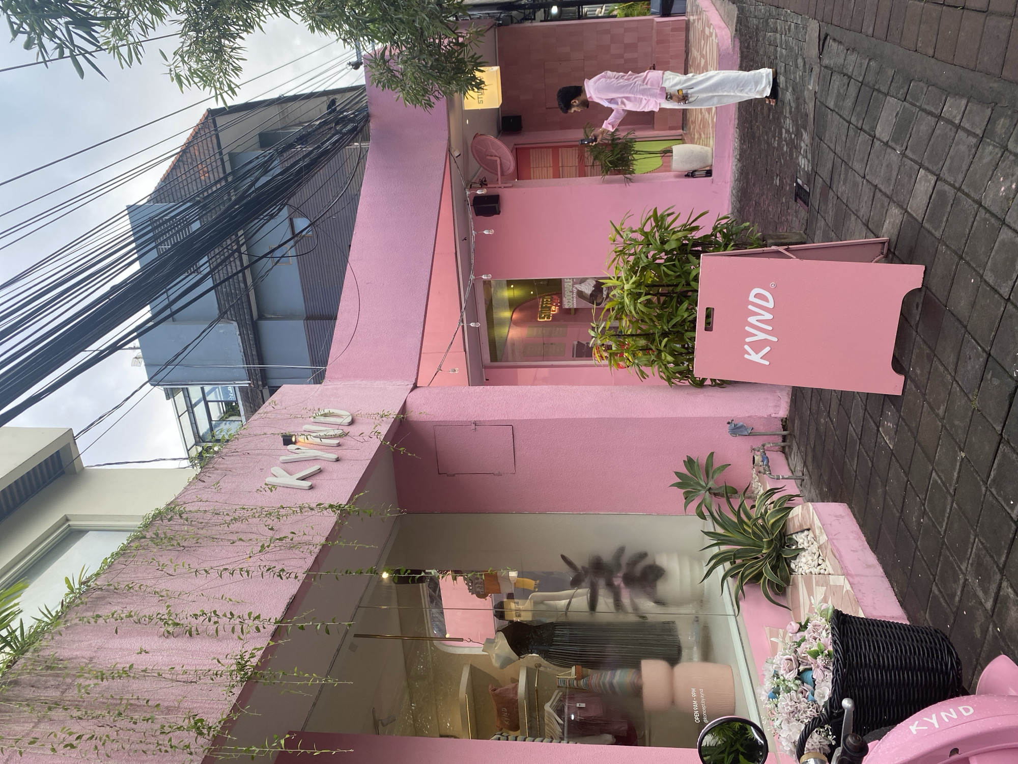 The image shows the exterior of a bright pink building in North Kuta, Indonesia. The building houses a shop called "KYND", as indicated by the signage on the facade and a freestanding signboard. The shop's front is decorated with plants and vines, and the display window showcases clothes and accessories. A man in a pink shirt and white pants is seen standing in the entrance area, looking at his phone. Power lines are visible overhead. The street is paved with gray bricks.