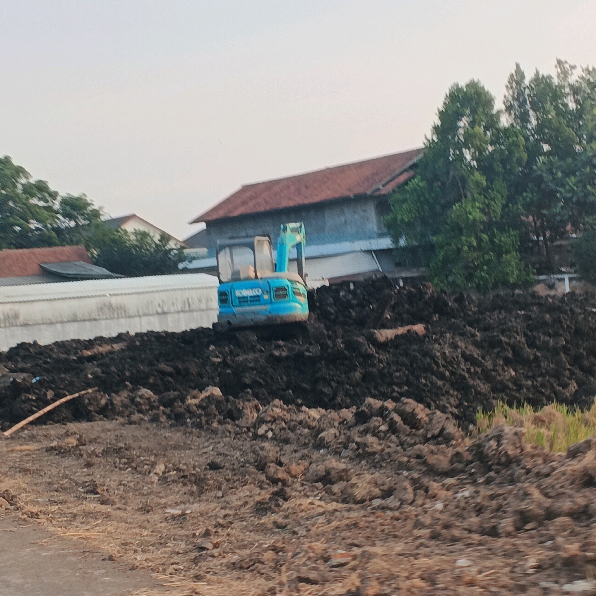 A bright blue excavator is parked on a large pile of dark soil. The excavator has the name "KOBELCO" written on its side. In the background, there are buildings with red-tiled roofs, a long white structure, and trees. The ground in the foreground is covered in dry grass and dirt. The sky is overcast. The scene appears to be a construction site in Gentasari, Indonesia.