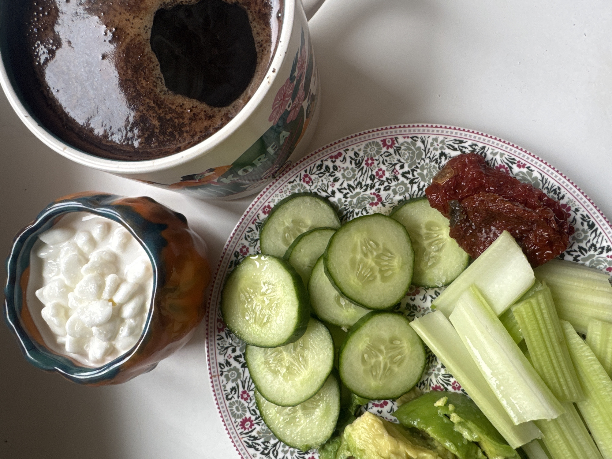 The image presents a top-down view of a light meal or snack arranged on a plain white surface, likely a table, in an indoor setting. The scene is illuminated by natural light, suggesting it is daytime.
In the upper left corner, a white ceramic mug, adorned with a vibrant floral and landscape design, contains a dark beverage, appearing to be coffee, with some foam and reflections on its surface. The word "KOREA" is partially visible on the side of the mug.
Below the mug, a small, organically shaped bowl with rich orange, brown, and teal glazes holds a portion of white, lumpy food, consistent with cottage cheese curds.
Dominating the right side of the frame is a round plate featuring an intricate floral and foliage pattern in grey, pink, and green around its rim. The plate holds a selection of fresh and prepared ingredients. Sliced green cucumbers are neatly arranged on the left side of the plate. To their right and towards the bottom, several light green celery sticks are visible. Chunks of green avocado are also present at the bottom right. In the upper right section of the plate, several pieces of reddish-brown, textured sun-dried tomatoes are displayed.
The arrangement suggests a healthy and varied meal or snack. The location context for this media file is Moscow, Russia.