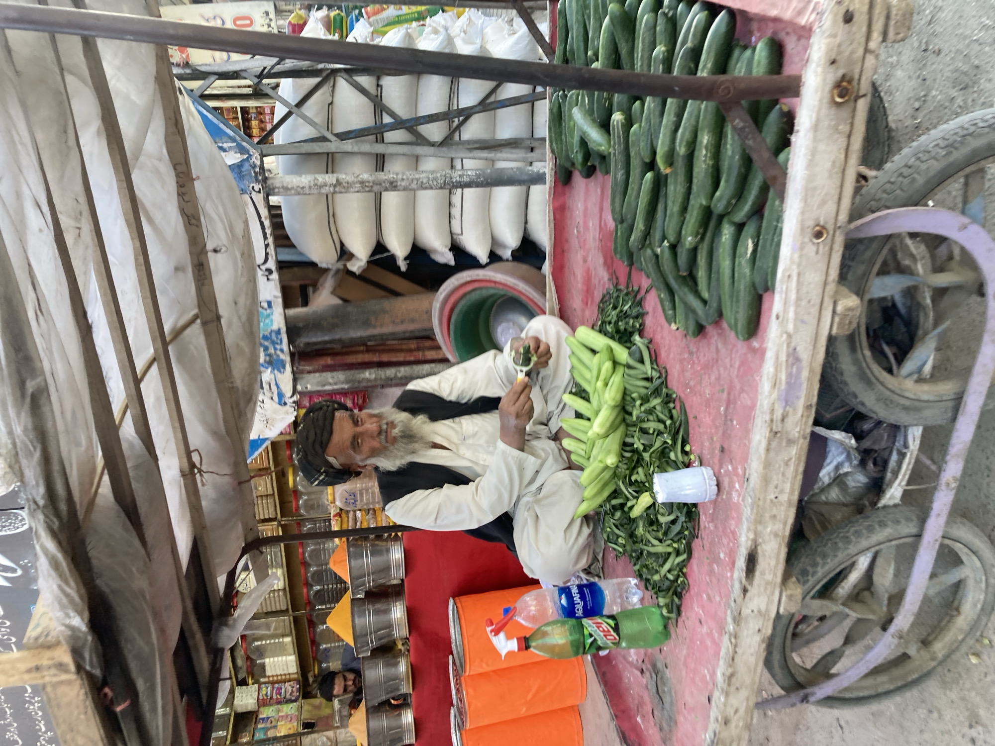 A street vendor with a long white beard wearing a turban and traditional clothing sits cross-legged on a red cart, in Quetta City Tehsil, Pakistan. The vendor is preparing produce, likely cucumbers, with a small tool. Large quantities of cucumbers are arranged in rows along the cart's side. Behind the vendor, the cart contains numerous white sacks stacked high. Rows of canned goods and other products are visible in the background, suggesting a nearby shop or store. The cart has two wheels and a makeshift canopy supported by a metal frame. Drinks are placed near the vendor.