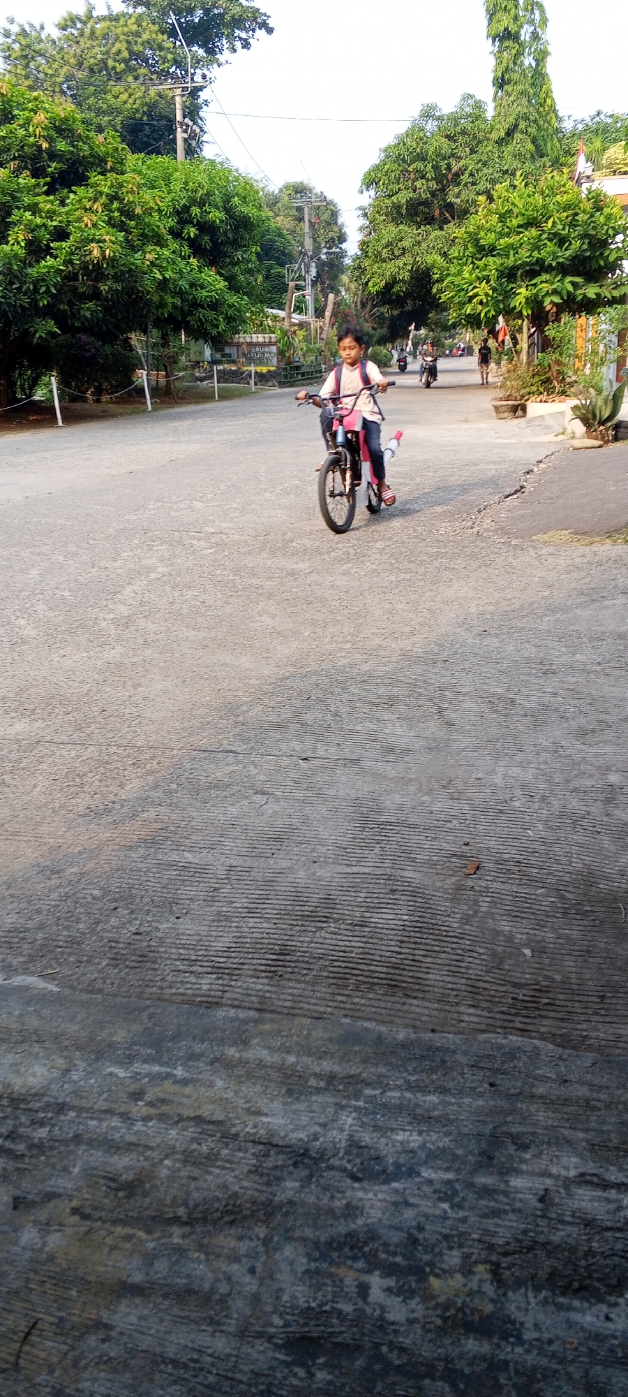 An outdoor, eye-level, full shot captures a young person riding a bicycle down a paved road in Depok, Indonesia. The person, with dark hair, is wearing a light-colored shirt, blue pants, and a backpack. The bicycle is red and black with a pink backpack attached. The road is textured, with visible parallel lines. Lush green trees line both sides of the road, with some houses and other people visible in the background. The sky is bright and overcast. The overall scene suggests a peaceful, everyday moment in a residential area.