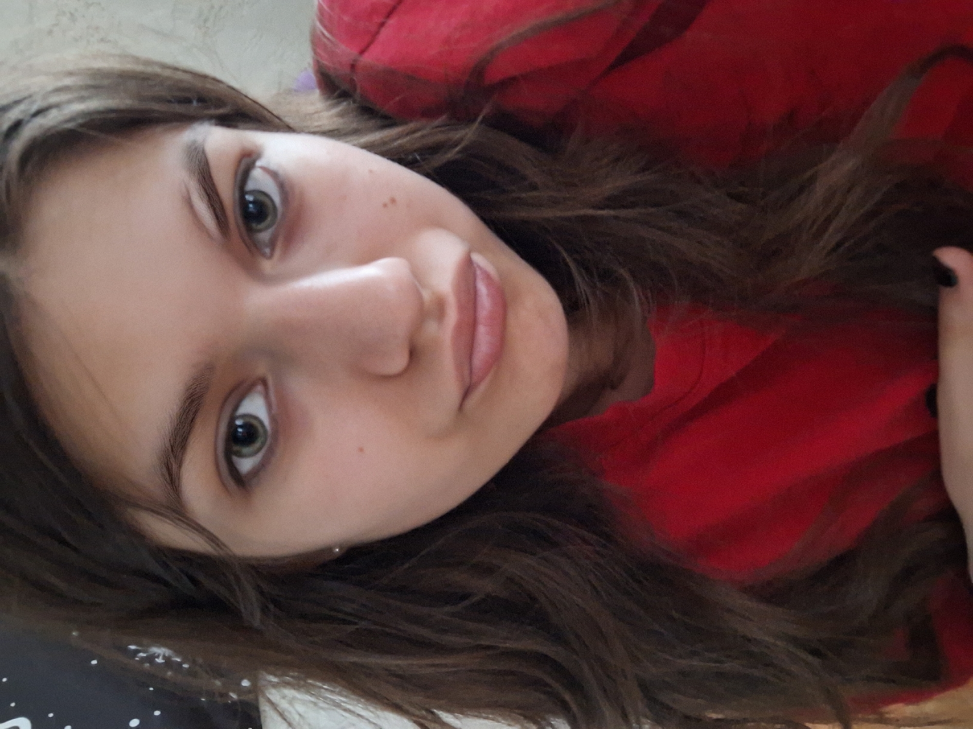 A young woman with light skin and brown hair is looking at the camera. She has light green eyes. She is wearing a red hoodie. The location is unknown.