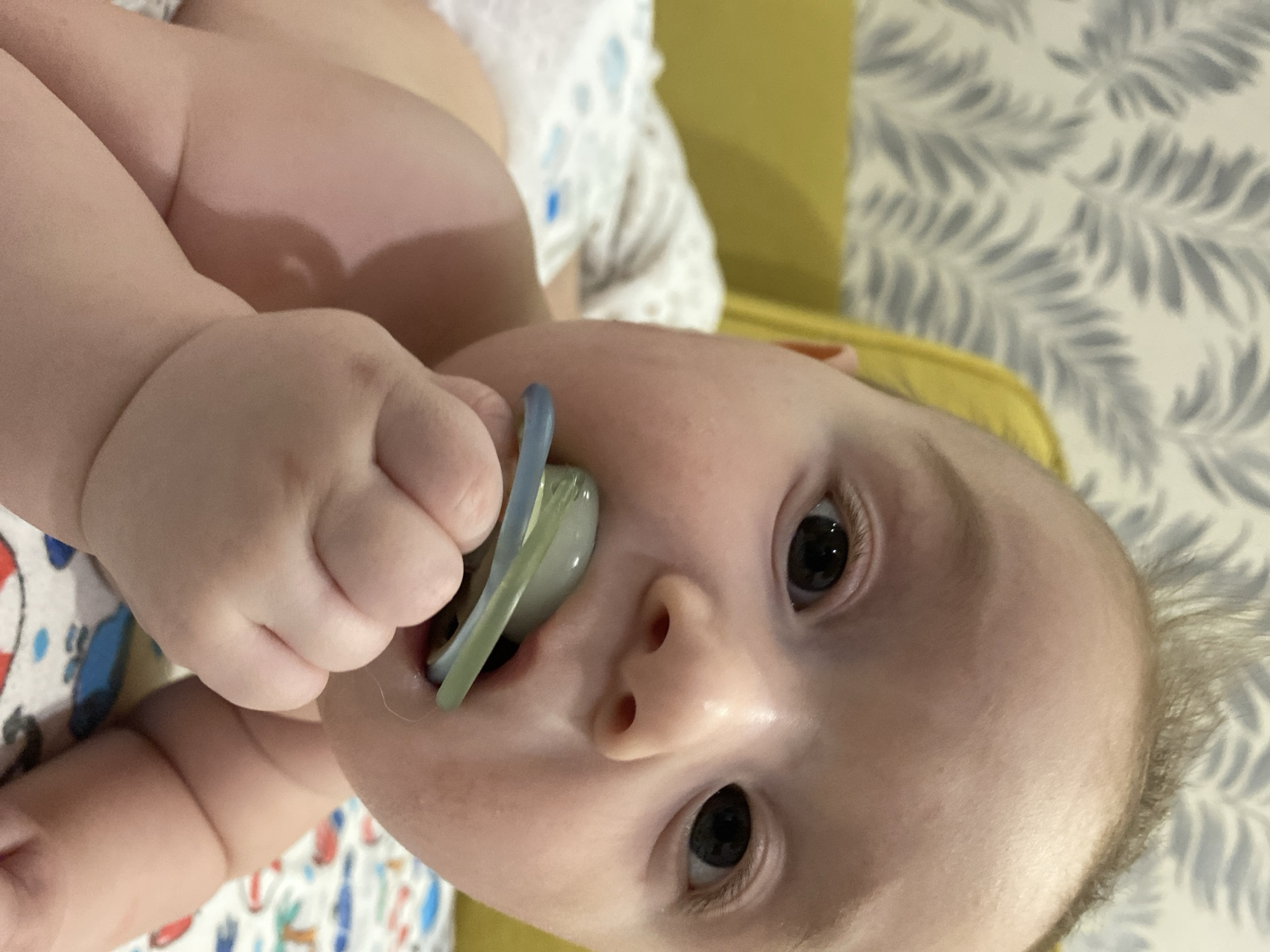 A close-up, slightly angled view captures a baby holding a pacifier in its mouth. The baby has fair skin, dark eyes, and a small nose. Its chubby hand firmly grasps the pacifier. The baby is wearing a diaper or clothing with a colorful, patterned print. In the background, there is a mustard-yellow surface and a wall covered in a light wallpaper with a grey leaf pattern. The city of Chaikovsky, Russia, is not discernible from the visual information provided in the image.