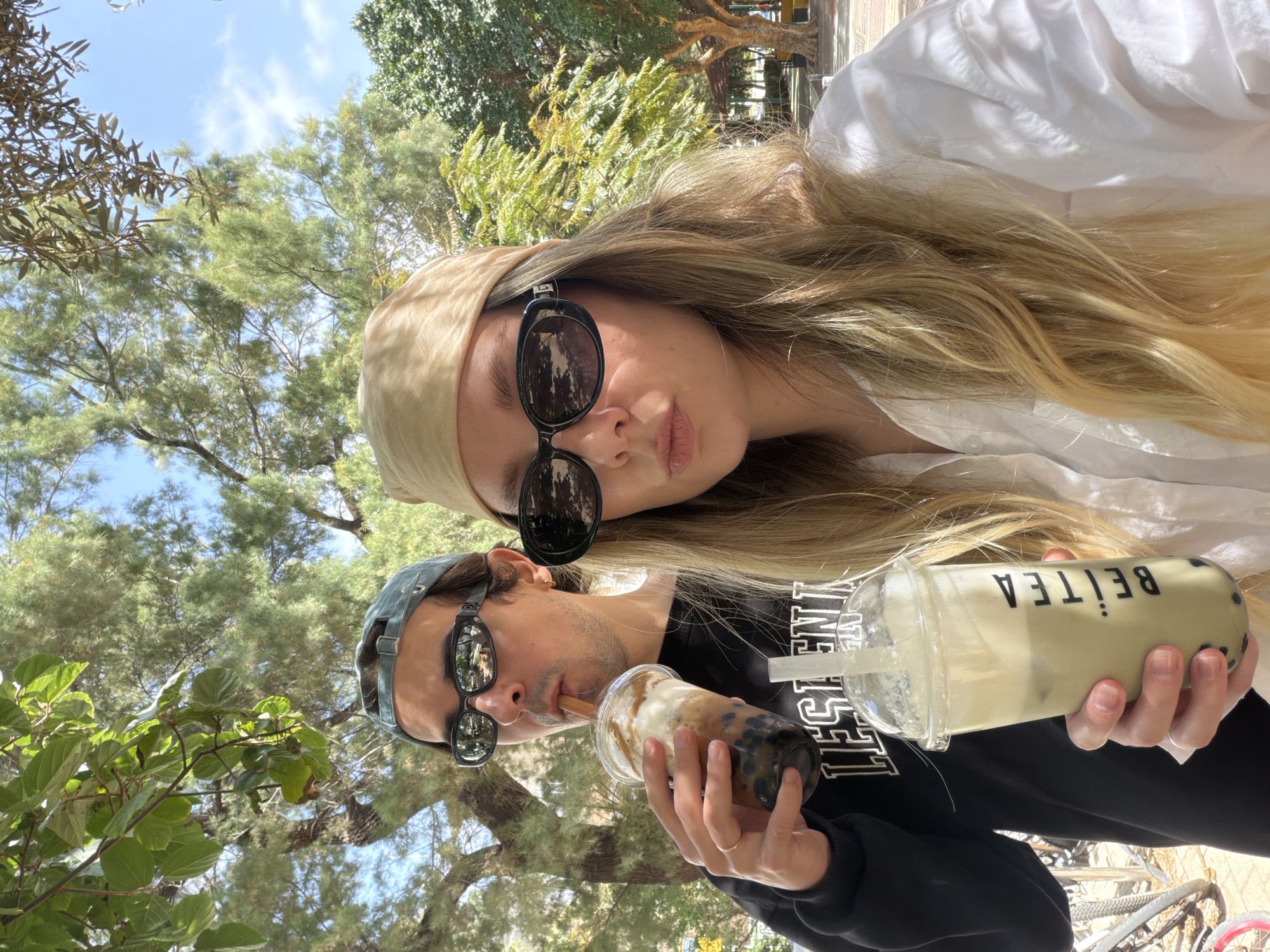 The image depicts two people taking a selfie while holding boba tea drinks. The woman on the top has long blonde hair, is wearing sunglasses and a light beige headscarf, and a white collared shirt. The man below her is wearing sunglasses and a baseball cap and drinking the boba through a straw. The woman holds a beverage branded "Beitea". The background features trees and a blue sky, likely indicating an outdoor setting. The environment suggests a possible park or recreational area in the city of Tel-Aviv, Israel.
