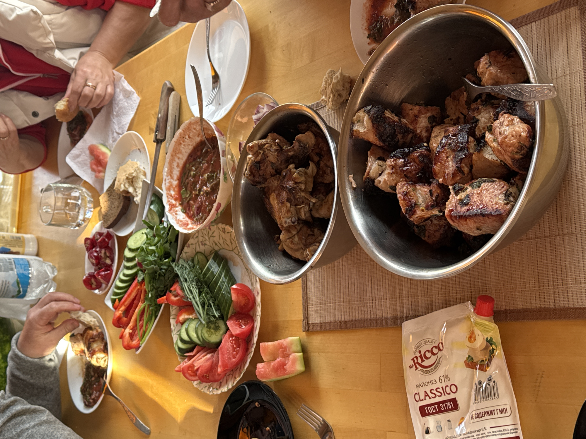 The image shows a tabletop laden with food, likely a meal being shared by several people. Visible on the table are two large metal bowls containing cooked meat, smaller bowls with what appears to be a tomato-based sauce and another with small red peppers, plates of sliced cucumbers, tomatoes, peppers, and herbs, and bread. There is also a packet of "Ricco Classico" mayonnaise, indicated as containing 61% fat. Parts of at least two individuals are visible at the table. One person is seen holding a piece of bread, and another is using a fork to eat from a plate. The food items suggest a meal is in progress. The mayonnaise packaging includes Russian text, possibly suggesting the meal is taking place in or with influences from Прикамский, Russia, though no definitive landmarks confirm the location.