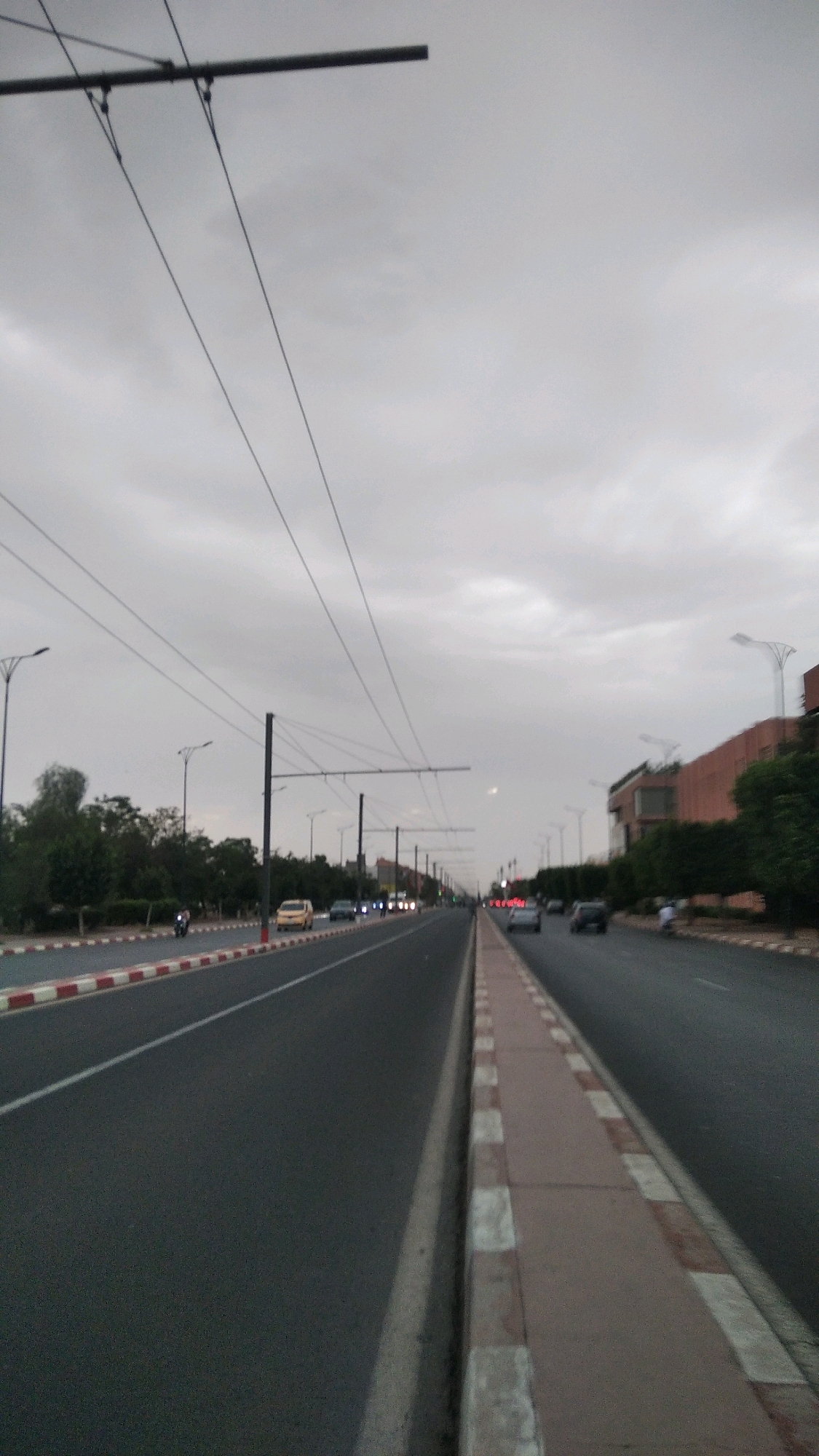 The image shows a wide, multi-lane road in a city. The sky is overcast, with grey clouds. There are overhead wires for what appears to be a public transportation system, such as a tram or trolleybus, stretching across the road. The road itself is asphalt, with distinct white and red striped curbs on the left side. On the right side, there is a pedestrian walkway with similar red and white patterned paving stones.
Several vehicles are visible on the road, including cars and a motorcycle. Some of the cars are moving, and their headlights are on, suggesting it might be dusk or a dimly lit area. Along the sides of the road, there are trees and some buildings. The buildings on the right appear to be modern, with some featuring brick facades. Streetlights are also visible, lining the road. The overall impression is that of a busy urban thoroughfare.