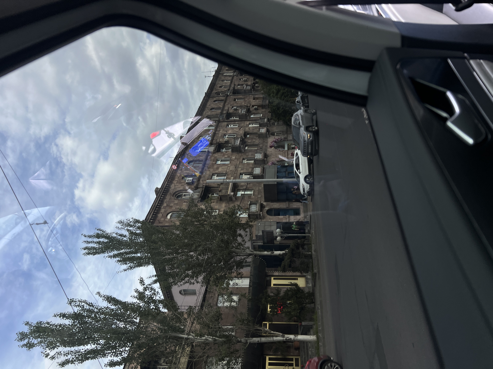 A medium-angle shot shows a street scene through a car window in Yerevan, Armenia. The window frame is visible on the left and right, with some reflections. On the right, there is a car door panel with a silver detail. Outside, there is a multi-story stone building with many windows and balconies, some of which have air conditioning units. A tree with bare branches is in the foreground. A street runs alongside the building, with parked cars, including a white vehicle and a gray one, visible. A few people are seen standing near the entrance of the building. A blue neon sign is attached to the building. The sky is partly cloudy.