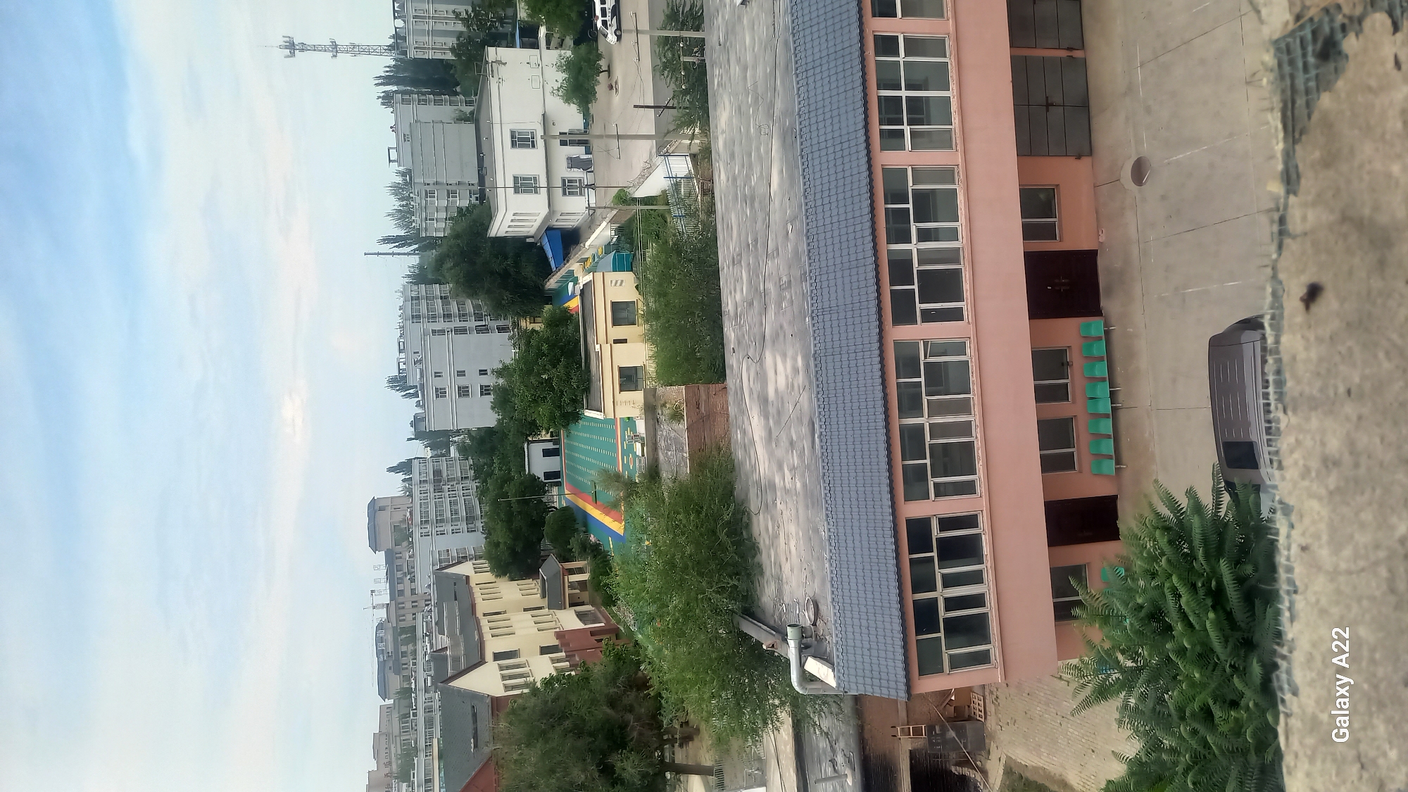 The image shows an aerial view of a building complex in Korgas City, China. In the foreground, a two-story pink building with a grey tiled roof is visible. It has numerous windows on both floors and several green benches are placed outside the ground floor. A dark-colored van is parked in the paved area in front of the building.
Behind this building, a colorful outdoor recreational area with a track and green spaces is visible. Further back, several residential buildings, characterized by their light-colored facades and numerous windows, are prominent. A telecommunications tower stands tall in the background, along with other urban infrastructure and a variety of trees. The sky is cloudy, suggesting it may be late afternoon or early evening. The overall scene depicts a residential and possibly educational or administrative area within the city.
