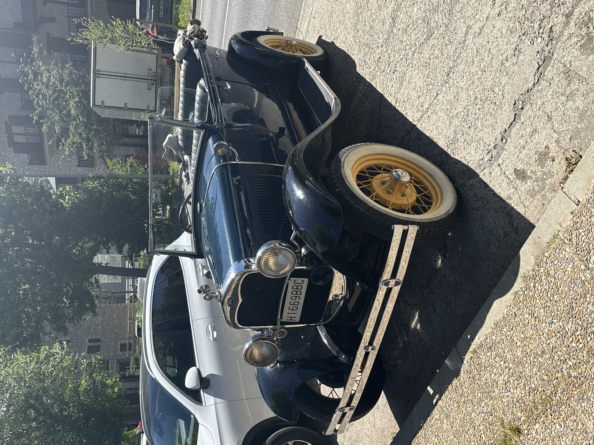 The image shows a vintage black Ford automobile with yellow spoke wheels and whitewall tires parked on the street in the city of Benasque, Spain. It is a convertible model with a chrome front bumper, headlights, and a license plate number H1669BBC. A modern white car is parked adjacent to the Ford. The street surface is asphalt, transitioning to a pebbled edge. Trees, buildings with stone facades, and a white truck are visible in the background, suggesting an urban or semi-urban setting. The lighting indicates it is daytime. No people are clearly visible.