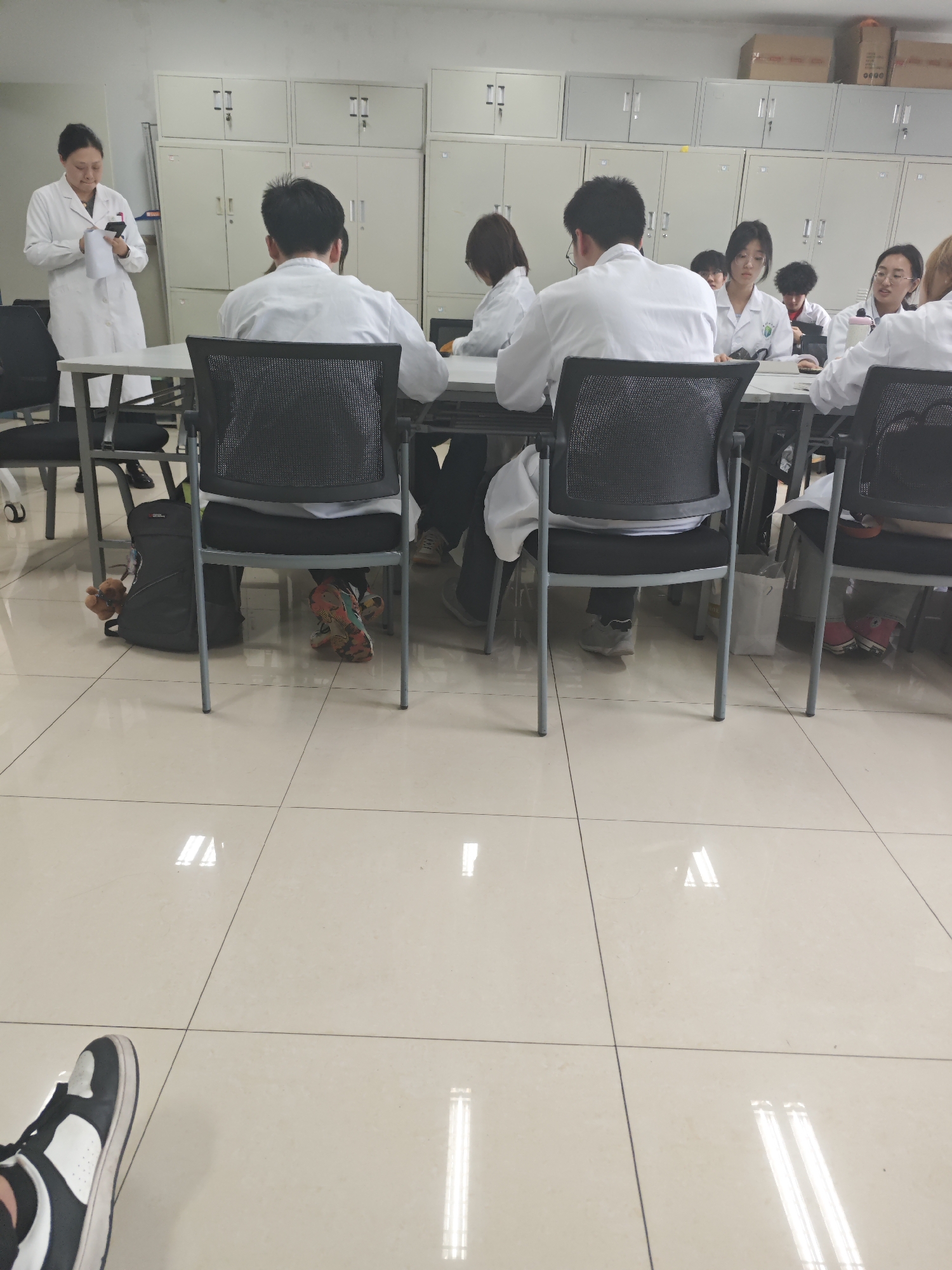 The image depicts an indoor scene within Yuzhong District, China, appearing to be a classroom or laboratory. Approximately ten to twelve individuals are visible. A woman in a white lab coat with dark hair pulled back stands on the left side of the frame, holding a white piece of paper in her left hand and a dark-colored smartphone in her right, seemingly interacting with both. Several individuals, also dressed in white lab coats, are seated at light-colored rectangular tables on chairs with dark mesh backs and metal frames. Two male individuals are prominently seated with their backs to the viewer in the foreground. Behind them, other participants are visible, including a woman with glasses and a green-and-white emblem on her lab coat. Footwear among the seated individuals varies, featuring colorful patterned sneakers, pink sneakers, and light grey sneakers. A black backpack with a brown stuffed animal keychain is positioned on the floor beneath a chair on the left. The background wall is lined with numerous light-colored metal storage lockers or cabinets, some of which have brown cardboard boxes stacked on top. The floor is composed of glossy, light-colored square tiles with dark grout lines, reflecting overhead rectangular light sources. The photographer's left foot, wearing a black and white sneaker, is partially visible in the lower-left corner of the frame.