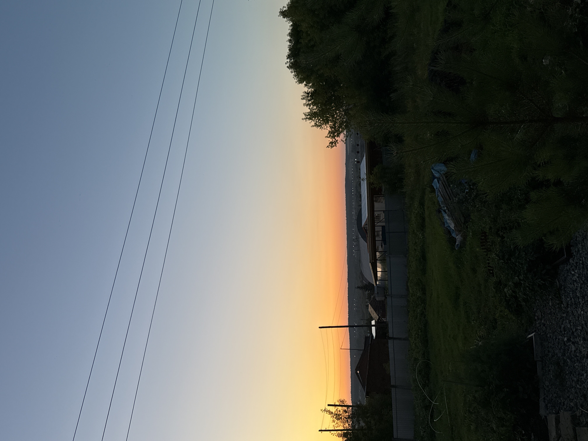 An outdoor, vertical, medium shot captures a sunset over a rural residential area in Malmyzh, Russia. The sky transitions from a soft, light blue at the top to a vibrant orange and yellow gradient towards the horizon. Three parallel power lines stretch horizontally across the upper portion of the image. In the middle ground, a row of houses with dark roofs and fences can be seen, partially obscured by trees and vegetation. The foreground consists of a dark, grassy area with a small pine tree and scattered debris. The overall atmosphere is tranquil and serene, typical of a rural sunset.