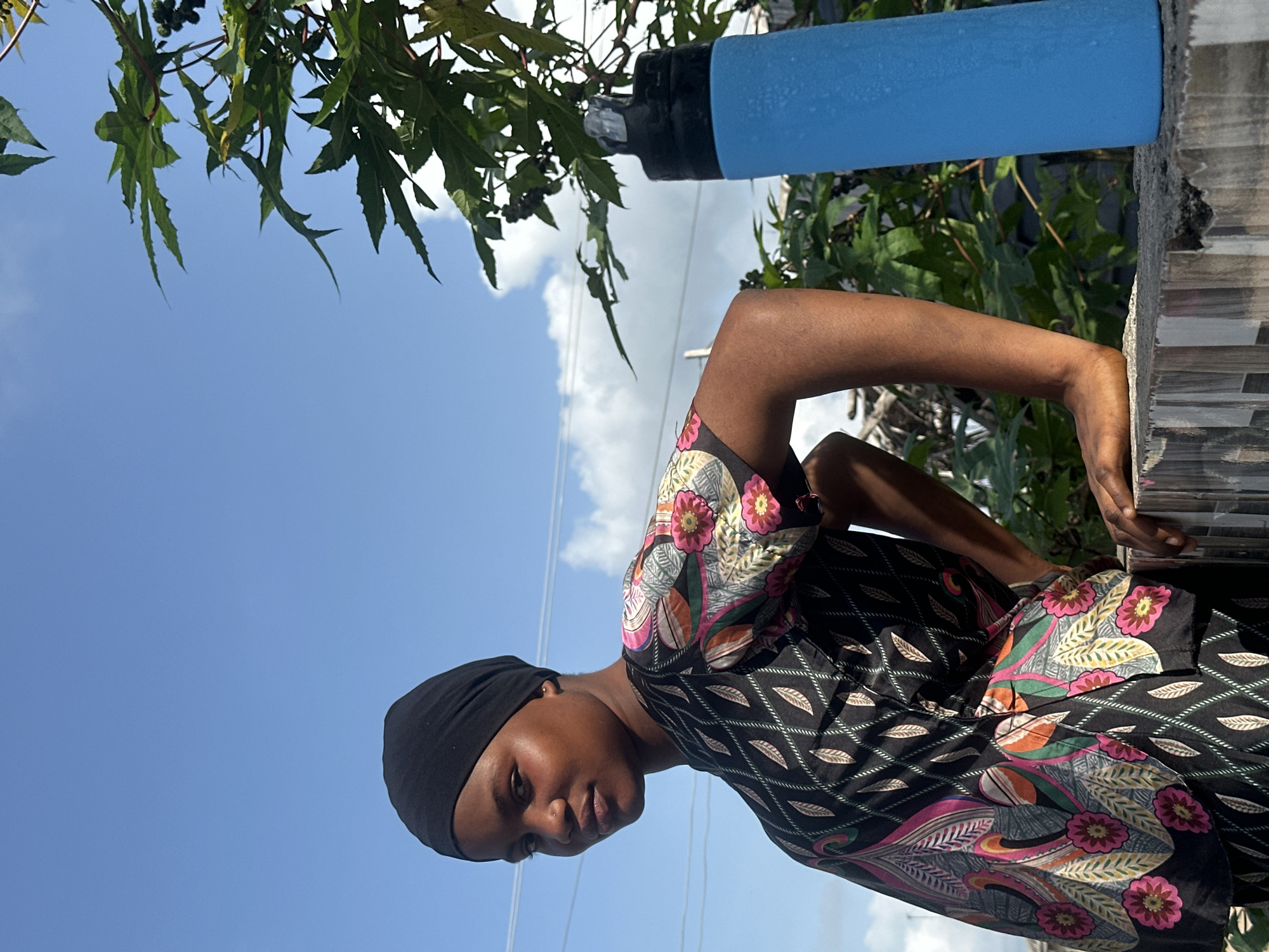 A medium shot captures a young person outdoors under a bright, clear blue sky with scattered white clouds in Badagry, Nigeria. The image is oriented 90 degrees clockwise from its likely original upright position.
The subject is a young person with dark skin, wearing a black head covering (possibly a hijab or headscarf) and a black top adorned with vibrant, colorful patterns of flowers and leaves in pink, orange, green, and light blue hues. They look towards the viewer with a neutral to slightly pensive expression. Their left arm is bent at the elbow, and their hand rests on a textured, possibly tiled or stone, wall or ledge visible on the right side of the frame.
The background primarily features the vast blue sky. Green foliage with distinctive jagged leaves, possibly from a tree or large plant, frames the upper portion of the scene, with some dark berry-like clusters visible within the leaves. A light blue cylindrical object, which appears to be a water bottle or insulated pipe with condensation or texture, is positioned in the upper right, partially obscured by the foliage and the wall. Thin, faint lines, possibly power lines, can be discerned against the sky. The scene is brightly illuminated by natural daylight, and there is no visible text.