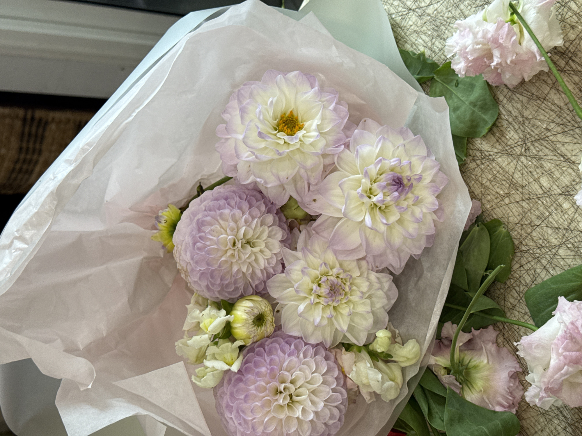 A bouquet of flowers, primarily dahlias and lisianthus, is presented. The dahlias are a mix of light purple and cream colors, with intricate petal formations. The lisianthus flowers are a soft pink and white, with delicate, ruffled petals. The bouquet is wrapped in light pink tissue paper, with some green foliage peeking out. The flowers are arranged on a textured surface with a crisscross pattern, possibly a table or mat. The background is softly blurred, with hints of a wicker object and a doorway. The image appears to be taken indoors, with natural light illuminating the subjects. There are no people or discernible actions in the scene.