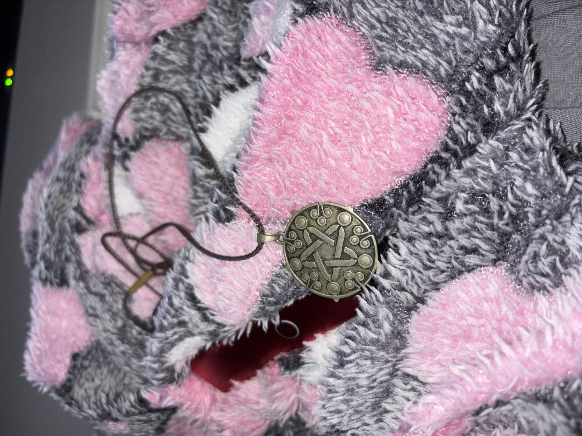 A close-up, overhead shot shows a bronze pentagram pendant hanging from a brown cord. The pendant rests on a checkered blanket with pink and gray fuzzy patches. The background is dark, with two green and orange lights visible at the top right. The scene is indoors, possibly in a bedroom. No specific location in Saint Petersburg, Russia, can be identified from the image.