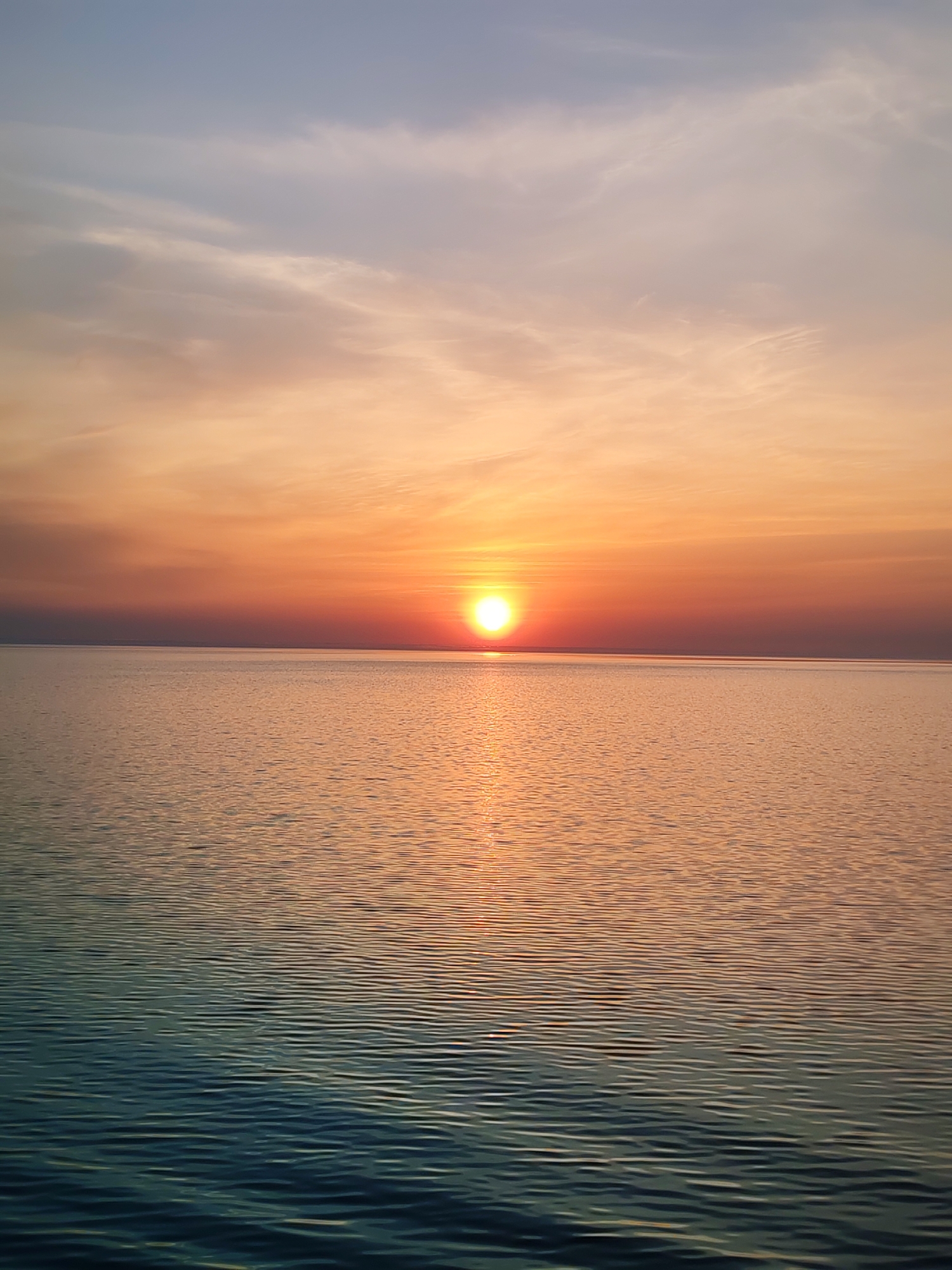 A wide-angle, eye-level shot captures a sunset over water. The sun, a bright orb, sits just above the horizon line, casting a shimmering reflection onto the water's surface. The sky transitions from shades of orange and red near the horizon to lighter blue tones higher up, with wispy clouds scattered throughout. The water displays small, gentle ripples, and its color graduates from a dark teal in the foreground to a golden hue near the horizon. There are no visible people or structures. Bol'shie Mkhi is not visibly represented and the environment is open water with a horizon at sunset.