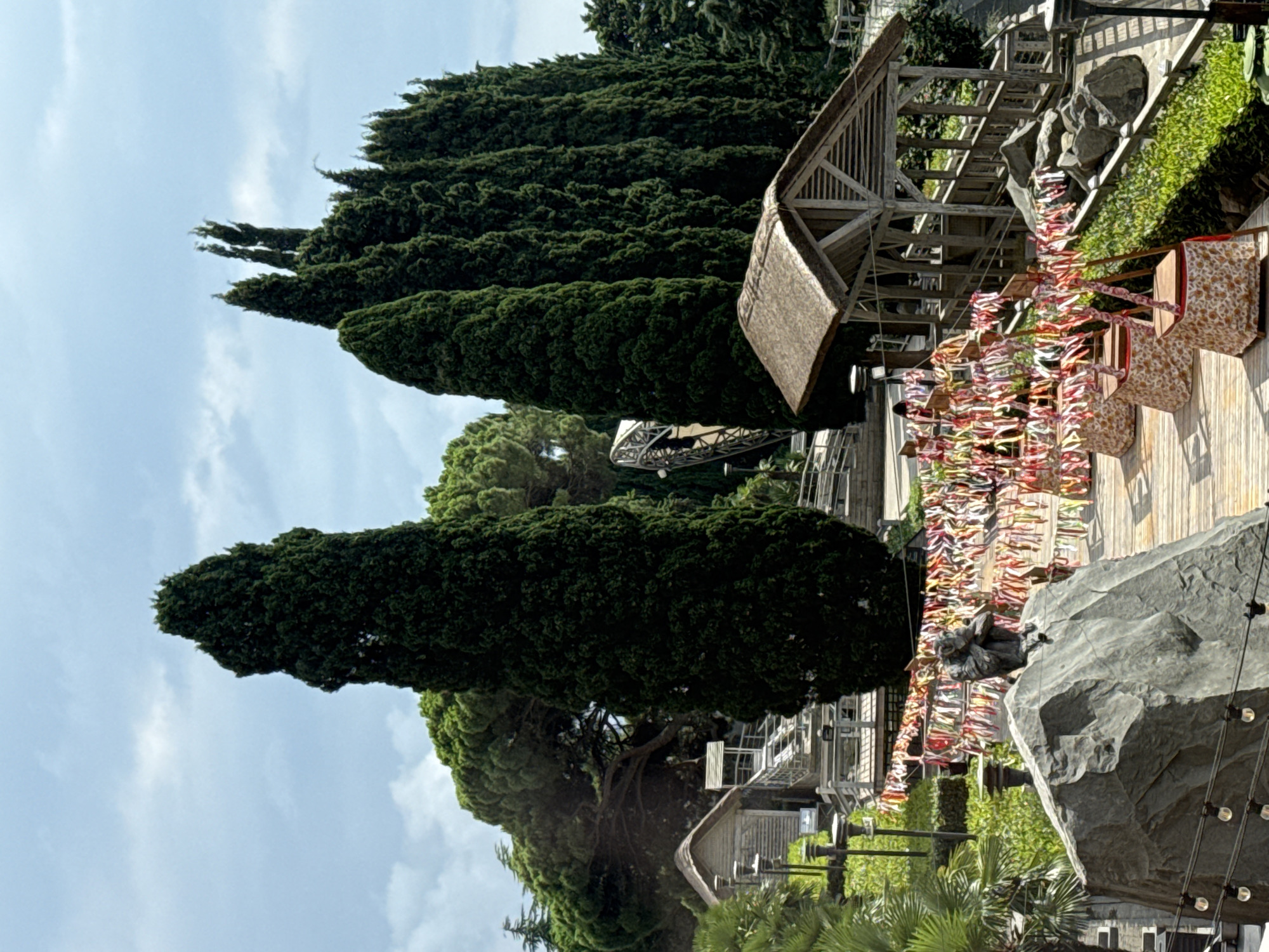 The image shows an outdoor scene featuring a pathway with wooden decking, lined with vibrant, colorful ribbons strung across the path. Several tall, dark green cypress trees dominate the background, with a large, rounded tree providing a contrasting silhouette against the sky. In the foreground, a large, grey rock is topped with a dark sculpture of a seated figure, possibly a monkey or ape. Further along the path, a rustic wooden structure with a thatched roof and open sides is visible, resembling a pavilion or shelter. Several chairs, covered with patterned fabric, are arranged near the structure. In the distance, more wooden structures and pathways are visible, along with lush green foliage and a cloudy sky. The scene appears to be a decorative outdoor area, possibly a park or a tourist attraction, located in the city of Yalta, Ukraine.