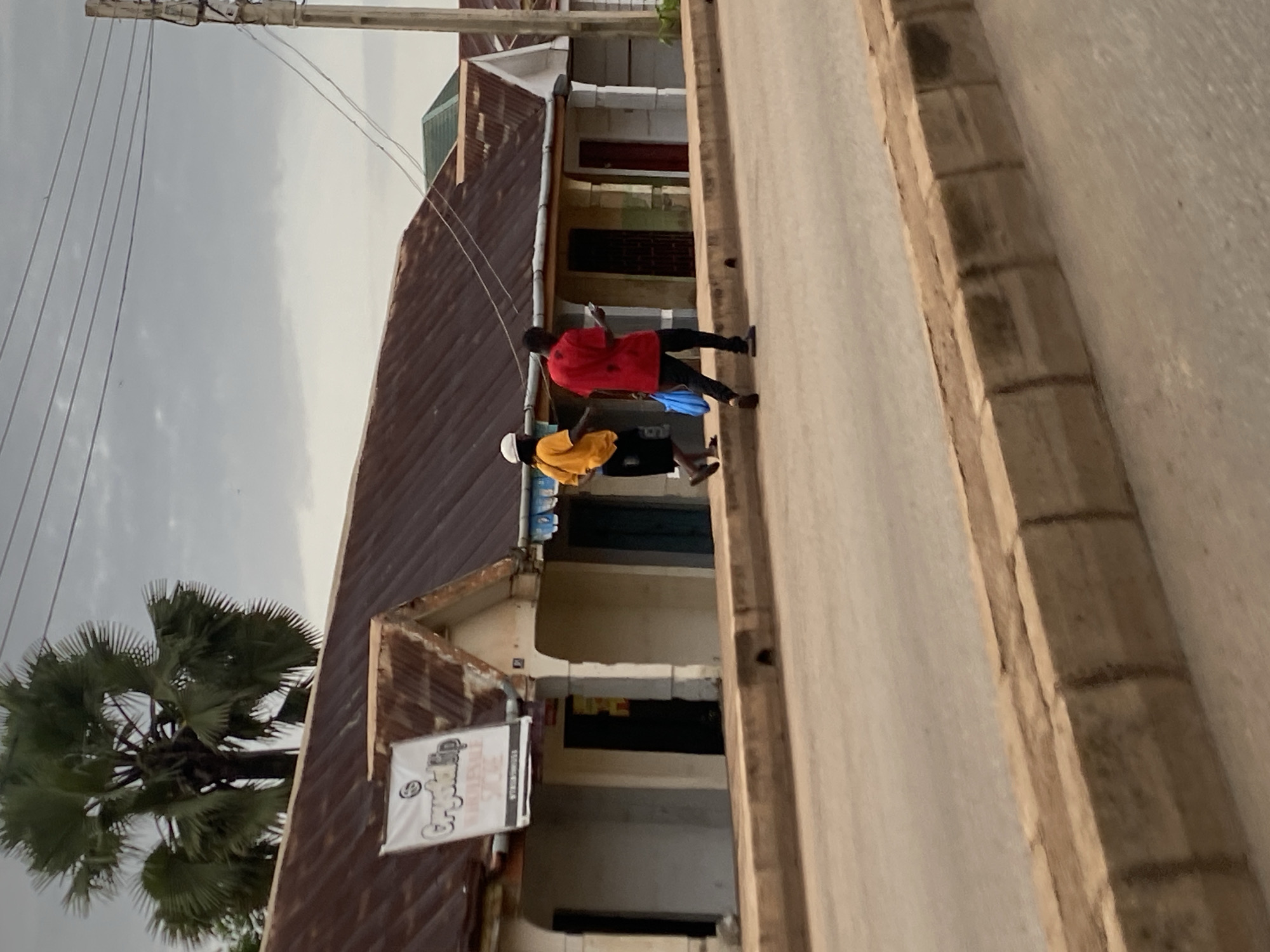 The image depicts a street scene in Barakin Lerawi, Nigeria, under an overcast sky. Two individuals are walking along a sidewalk in front of a row of buildings. The person on the left wears a yellow t-shirt and black shorts, with a white cap. The person on the right wears a red t-shirt and black trousers, carrying a blue plastic bag and looking at a mobile phone. The buildings have corrugated metal roofs and storefronts. A palm tree is visible behind the buildings. The sidewalk is bordered by a concrete curb, and a paved road runs in front of it. Power lines and a utility pole are present in the background.