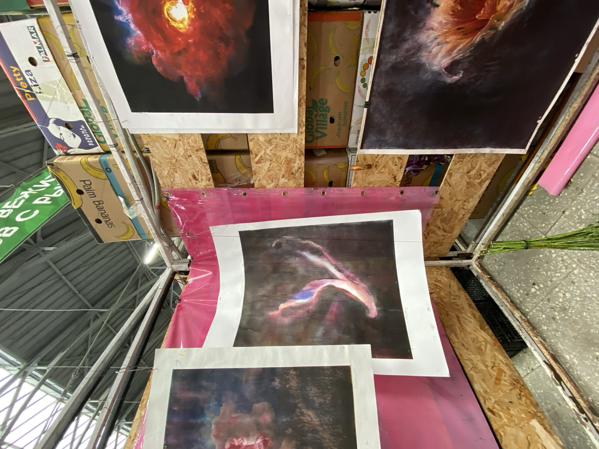The image shows an indoor market stall displaying various pieces of art. The artwork consists of several large prints, some framed and some unframed, featuring abstract and nebula-like imagery with vibrant colors. The prints are hung on a temporary structure made of plywood and metal framing, with some draped over pink plastic sheeting. In the background, stacked cardboard boxes, some with banana imagery and branding such as "Pretty Liza" and "Global Village Premium bananas," suggest a market environment, possibly a fruit market repurposed for art. Above, a metal ceiling structure with exposed beams and a glass roof indicates a large, covered space. The flooring appears to be concrete with visible aggregate. A bunch of green stems is visible in the foreground, tied together with string. There are no people visible in the frame. The lighting suggests an overhead light source. The scene is consistent with a pop-up art exhibition or a stall in a large, indoor market, potentially in the city of Saint Petersburg, Russia, given the context provided.