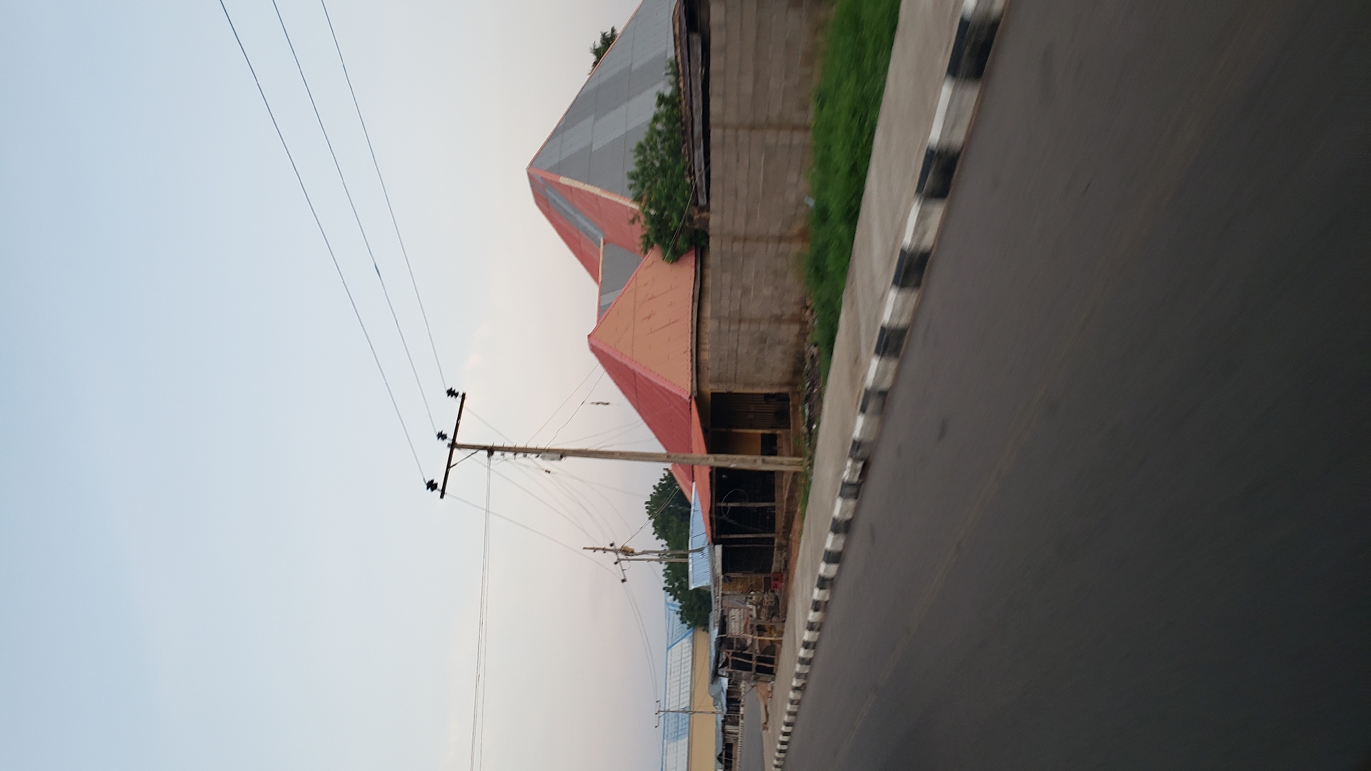 The image depicts a roadside scene in Jalingo, Nigeria, under a pale sky at what appears to be dawn or dusk. A paved road occupies the foreground, with a distinct black-and-white striped curb separating it from a sidewalk. Beyond the sidewalk, a row of modest structures lines the street. One building features a striking red, pyramidal roof, adjacent to another structure with a similarly colored roof, though it appears smaller and partly obscured. Further down the street, a larger, more complex roof structure is visible, its upper section a gray and white geometric pattern, suggesting a more significant building or complex. Electrical poles with numerous wires crisscross the sky above these buildings, indicating a populated area with power infrastructure. Some of the smaller structures appear to be shops or stalls, with open fronts and rudimentary construction. The overall impression is one of a developing urban environment, with a mix of residential and commercial buildings. There are no people visible in the image.